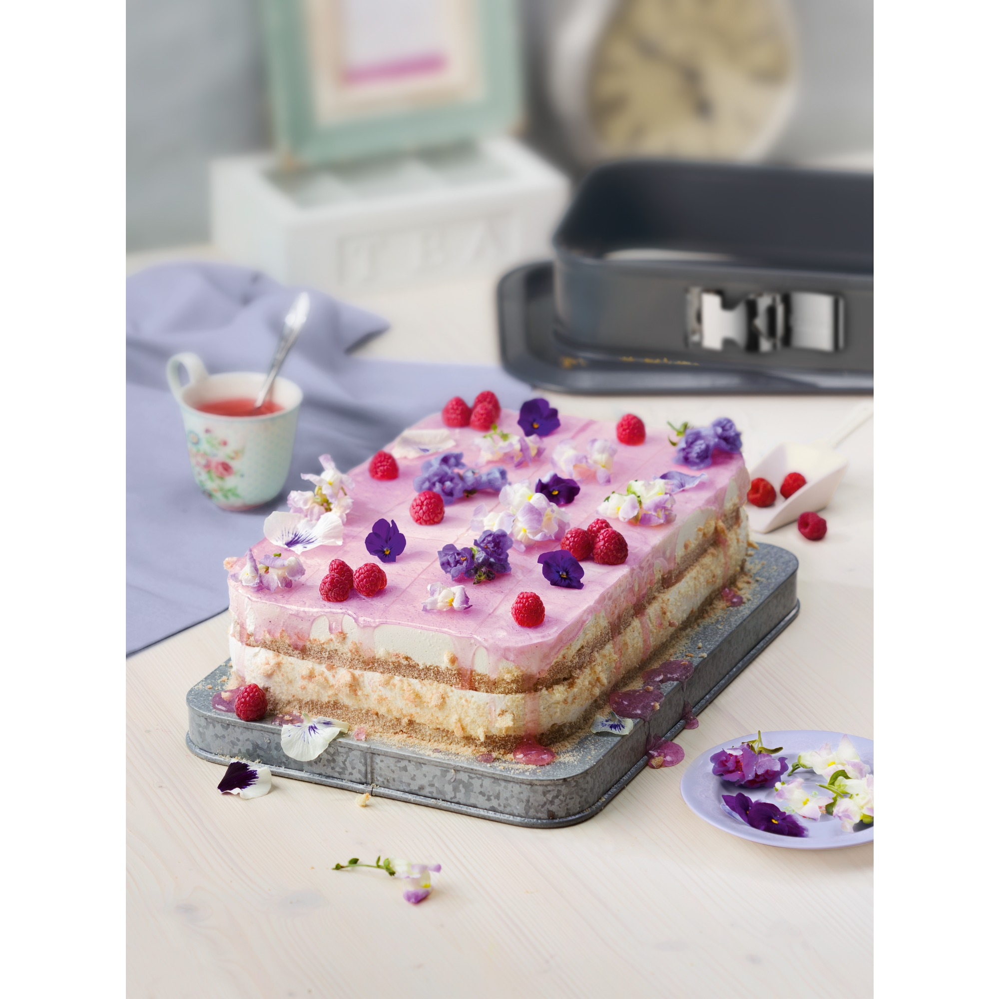 Un gâteau à étages avec glaçage rose, framboises et fleurs sur un plateau, avec une tasse de thé en arrière-plan.