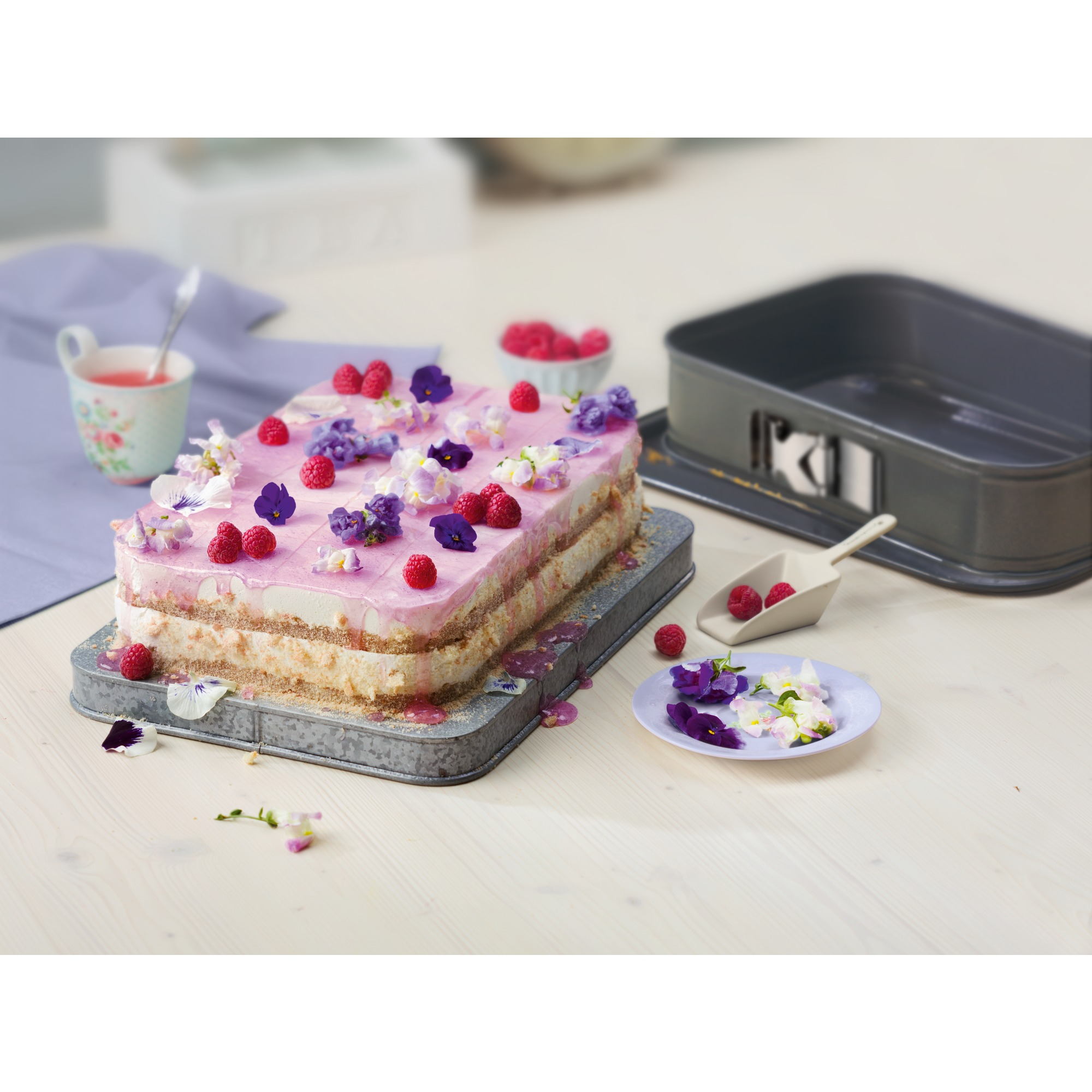 Un gâteau à étages avec glaçage rose est décoré de framboises fraîches et de fleurs comestibles sur une table.