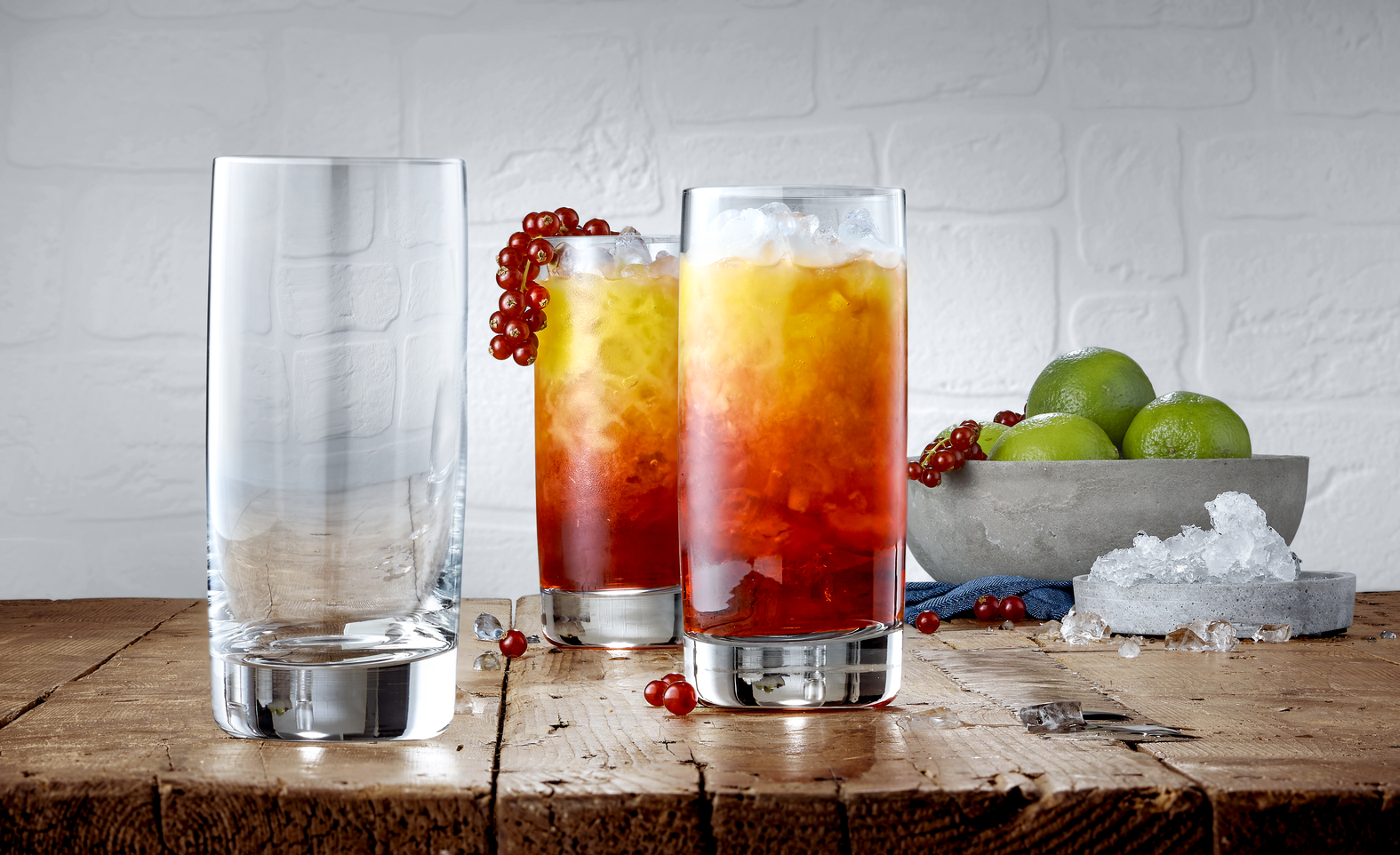 Drei hohe Gläser auf Holztisch, zwei mit farbigem Cocktail und Beeren, daneben Limetten in Schale.