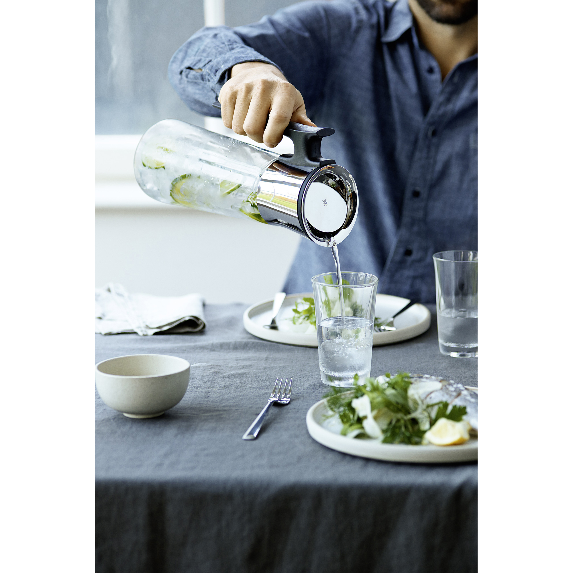 Un uomo con una camicia blu versa acqua da una caraffa di vetro con lime in un bicchiere.