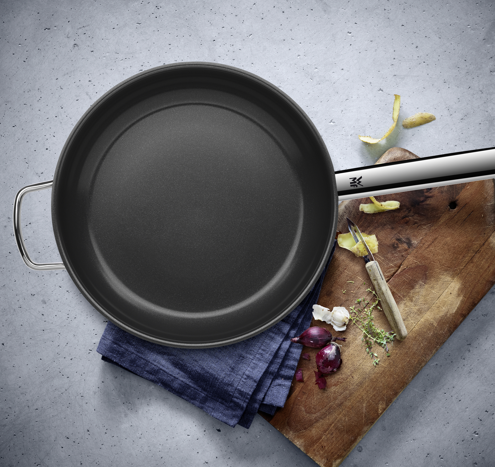 Eine schwarze Pfanne liegt auf einem blauen Tuch und Holzbrett mit Sparschäler, Kräutern und Zwiebelschalen.