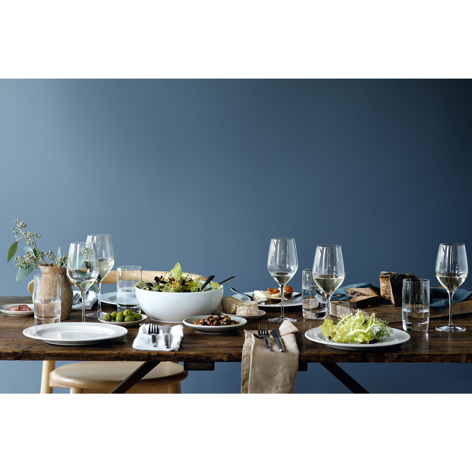 Ein rustikaler Holztisch mit Salaten, Brot, Oliven und Weingläsern vor einer dunkelblauen Wand.