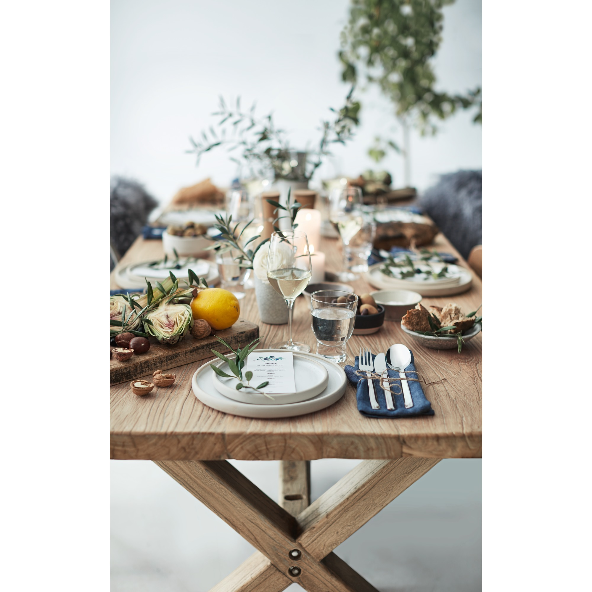 Ein rustikaler Holztisch ist gedeckt mit Tellern, Gläsern, Olivenzweigen, einer Zitrone und Artischocken.