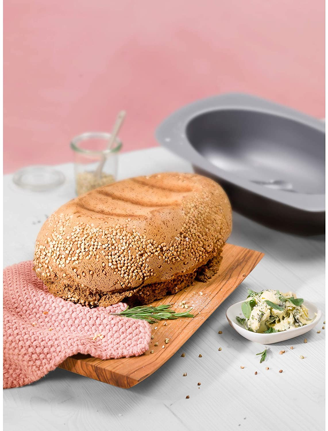 Ein Laib Brot mit Buchweizen auf einem Holzbrett, daneben Kräuterbutter, eine Backform und ein rosa Handtuch.