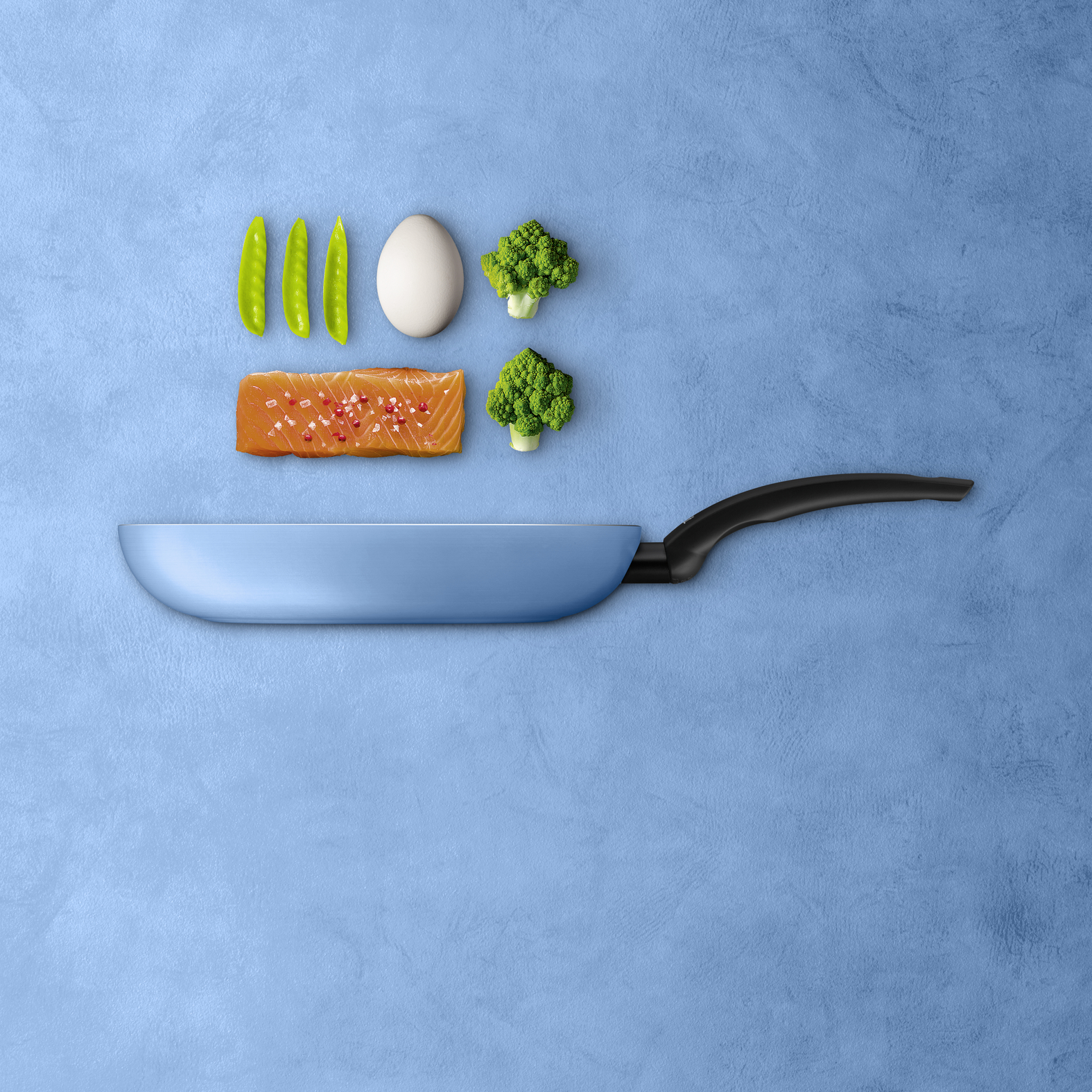 Blaue Pfanne auf blauem Hintergrund mit rohem Lachs, Ei, Erbsen und Brokkoli.