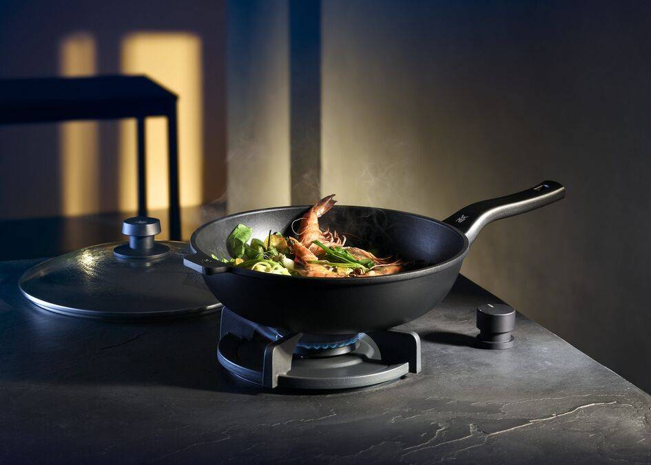 Eine schwarze Wok-Pfanne mit Garnelen und Gemüse brutzelt auf einem Gasherd in einer modernen Küche.