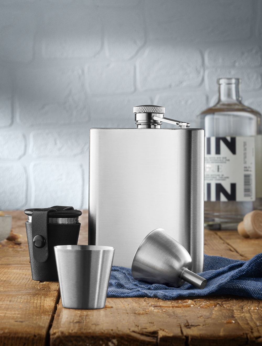 Edelstahlflasche, Trichter und Becher auf einem Holztisch mit blauer Serviette und Flasche im Hintergrund.