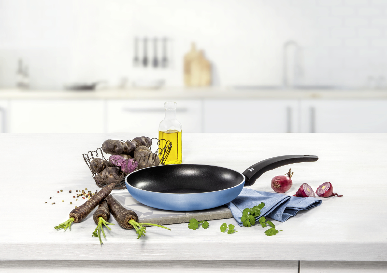 Blaue Pfanne auf weißem Tisch mit Öl, Kartoffeln, lila Karotten, Zwiebeln und Kräutern.