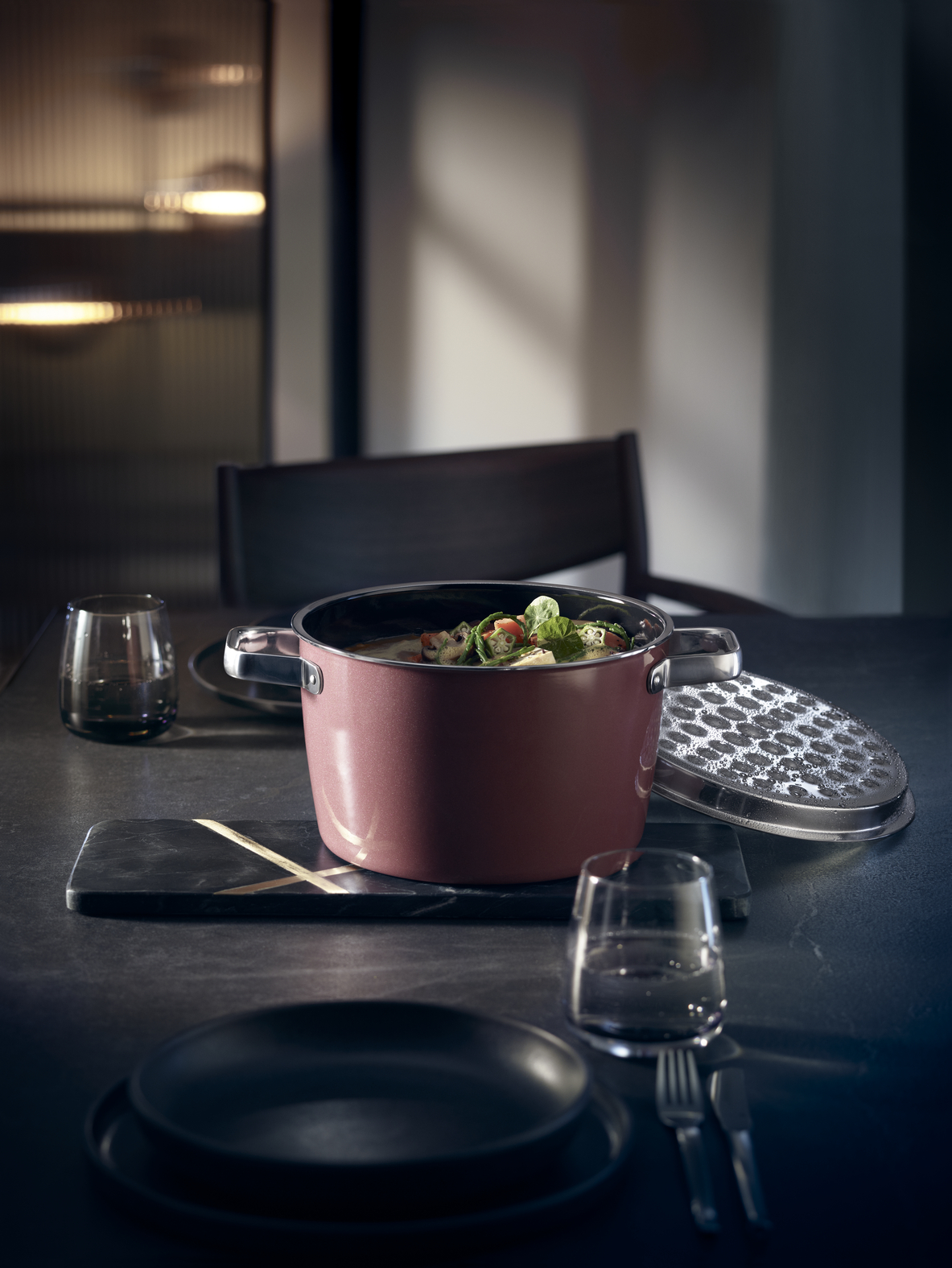 Ein rosafarbener Topf mit dampfender Suppe steht auf einem dunklen Tisch, begleitet von Gläsern und Tellern.