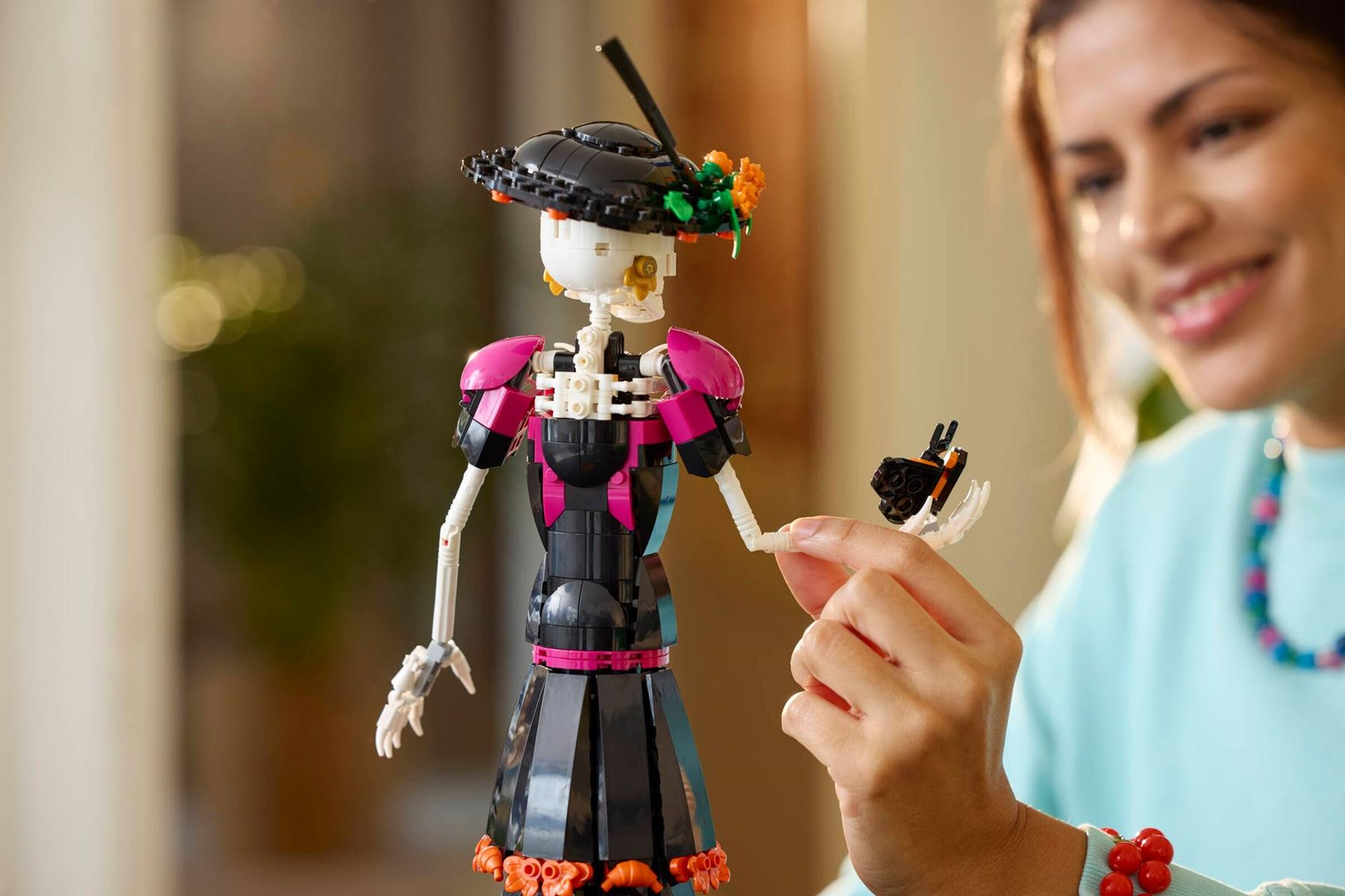 Una donna sorridente tiene una figura di scheletro Lego con un vestito nero e rosa e un cappello.