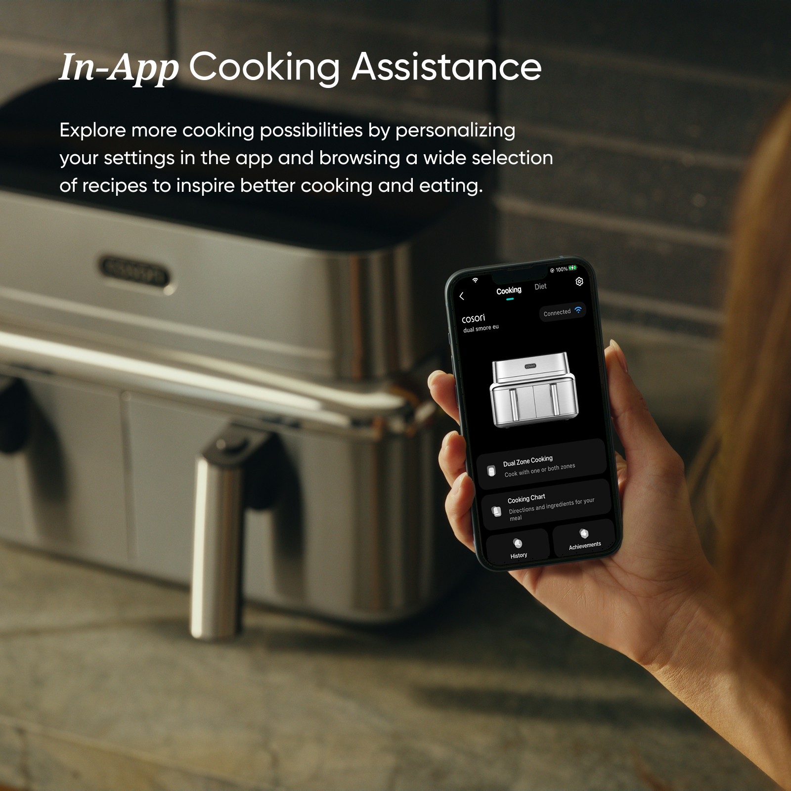 Una persona sostiene un smartphone con una aplicación de cocina y una freidora de aire de acero al fondo.
