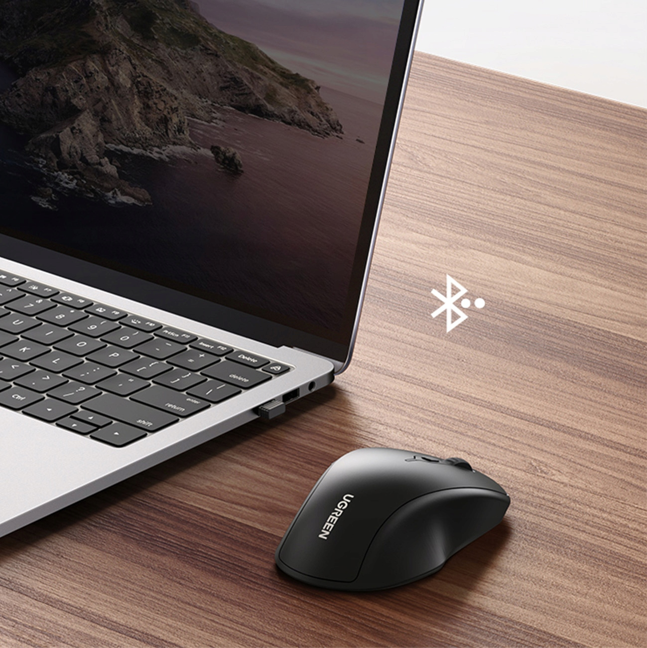 Szara klawiatura laptopa i czarna mysz bezprzewodowa na drewnianym biurku obok ikony Bluetooth.