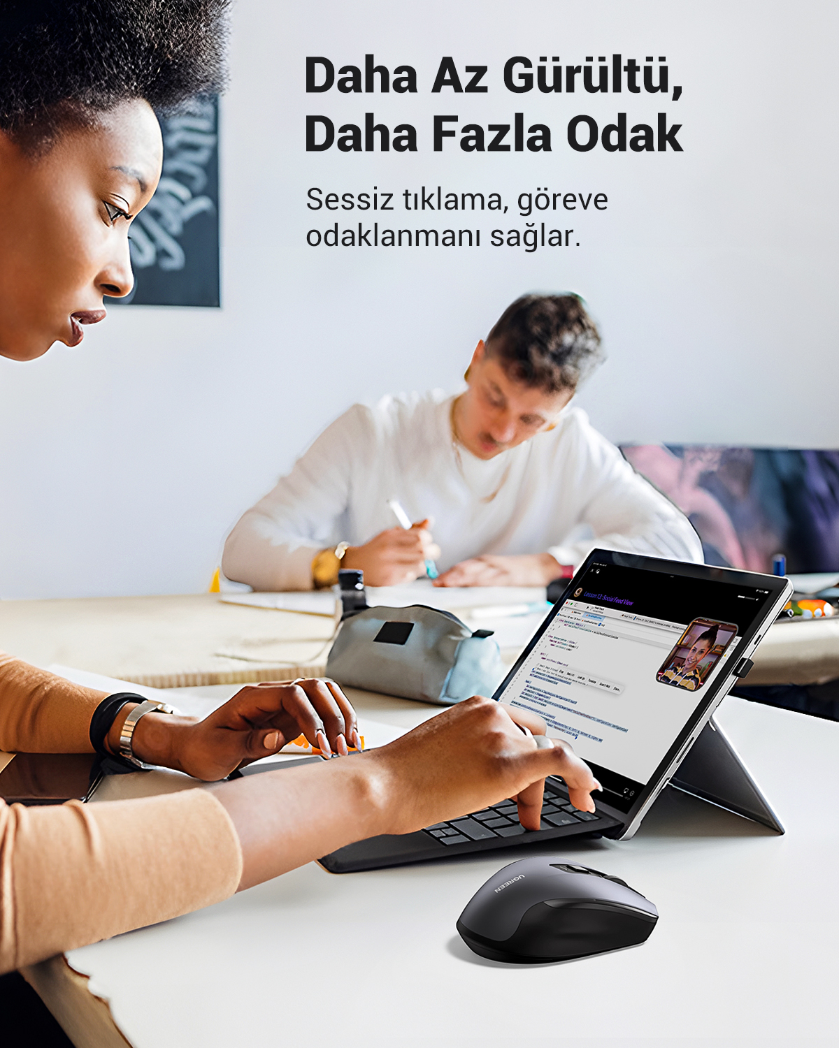 Bir kadın klavyeli tablette yazıyor, yanında bir fare, arka planda yazan başka bir kişi.
