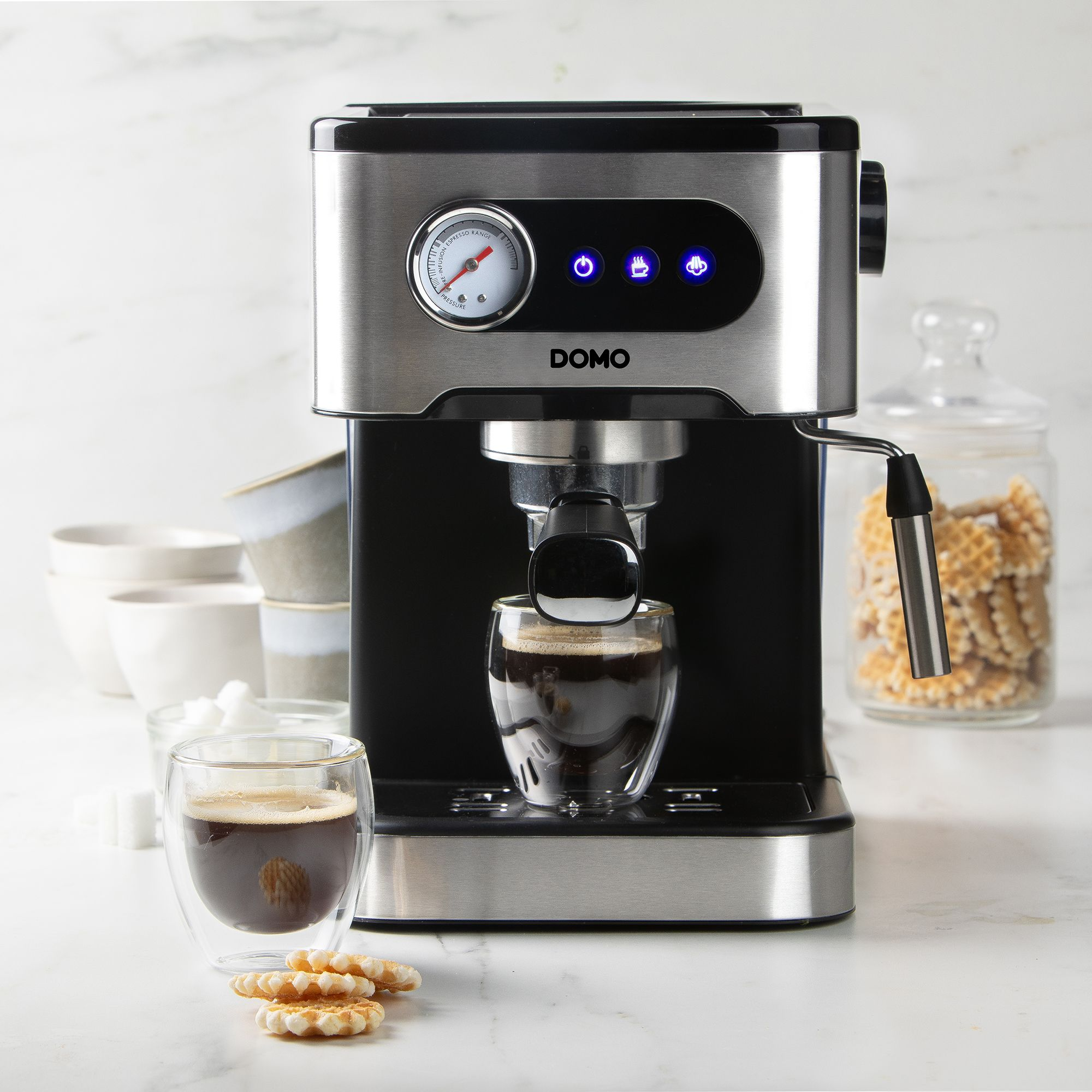 Una macchina da caffè Domo argento e nera prepara il caffè, con due bicchieri di caffè, biscotti e un barattolo sul bancone.