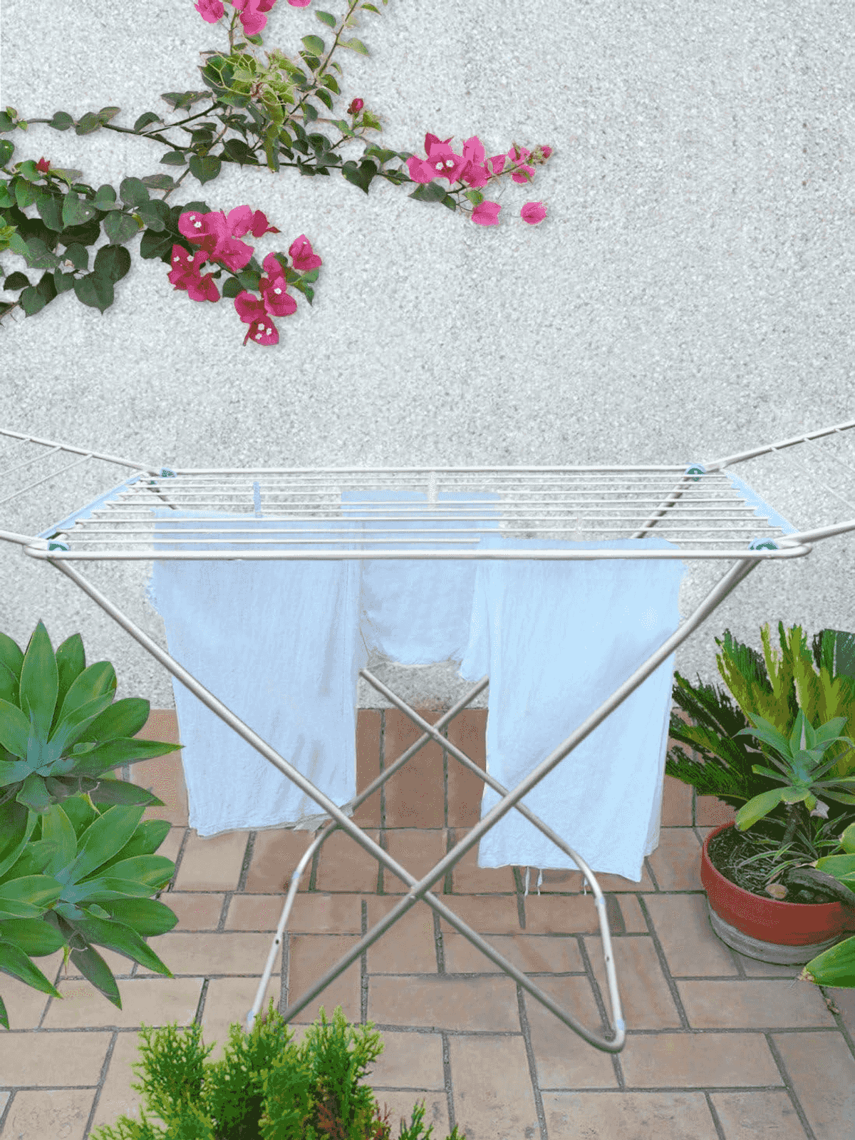 Un tendedero blanco con ropa blanca está en un patio de baldosas, rodeado de plantas en macetas y un muro con flores rosas.