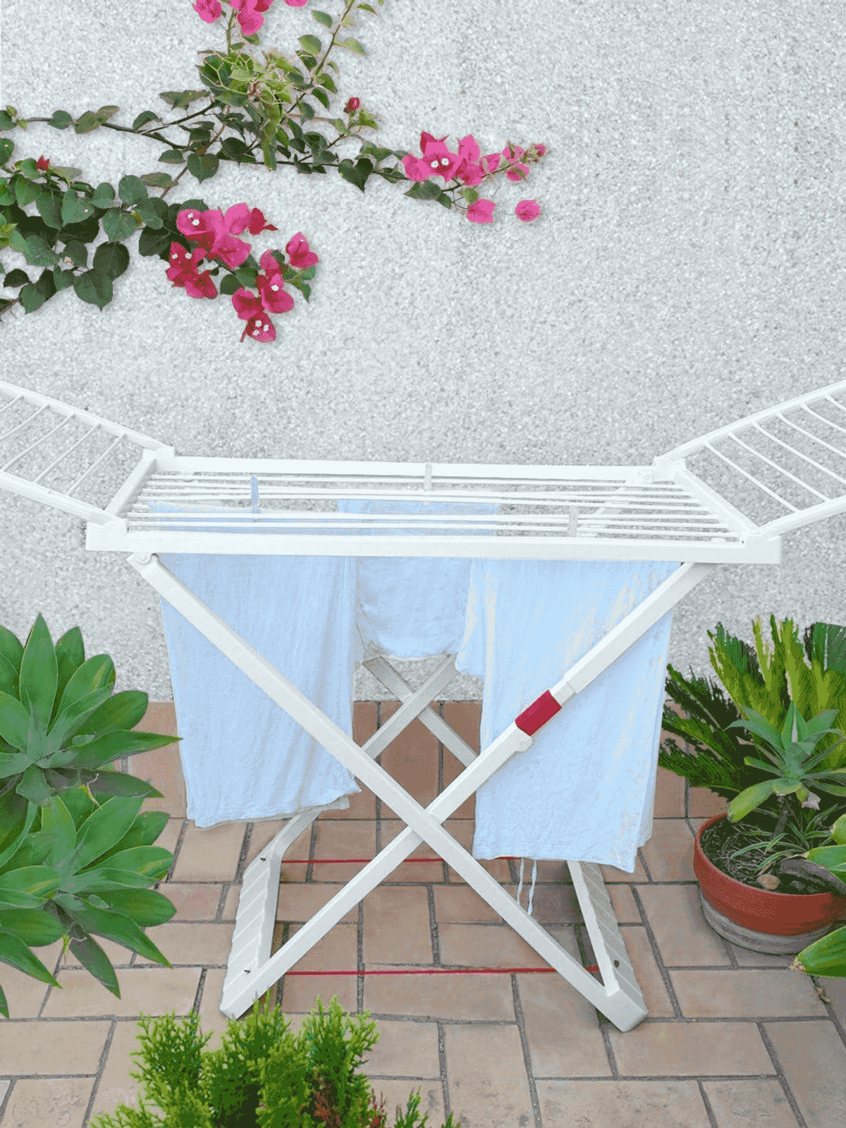 Un tendedero blanco plegable con ropa tendida sobre un patio embaldosado, bordeado de plantas verdes y una pared con textura.