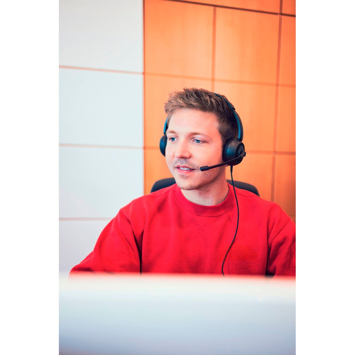 Un jeune homme en pull rouge et casque est assis devant un mur blanc et orange.