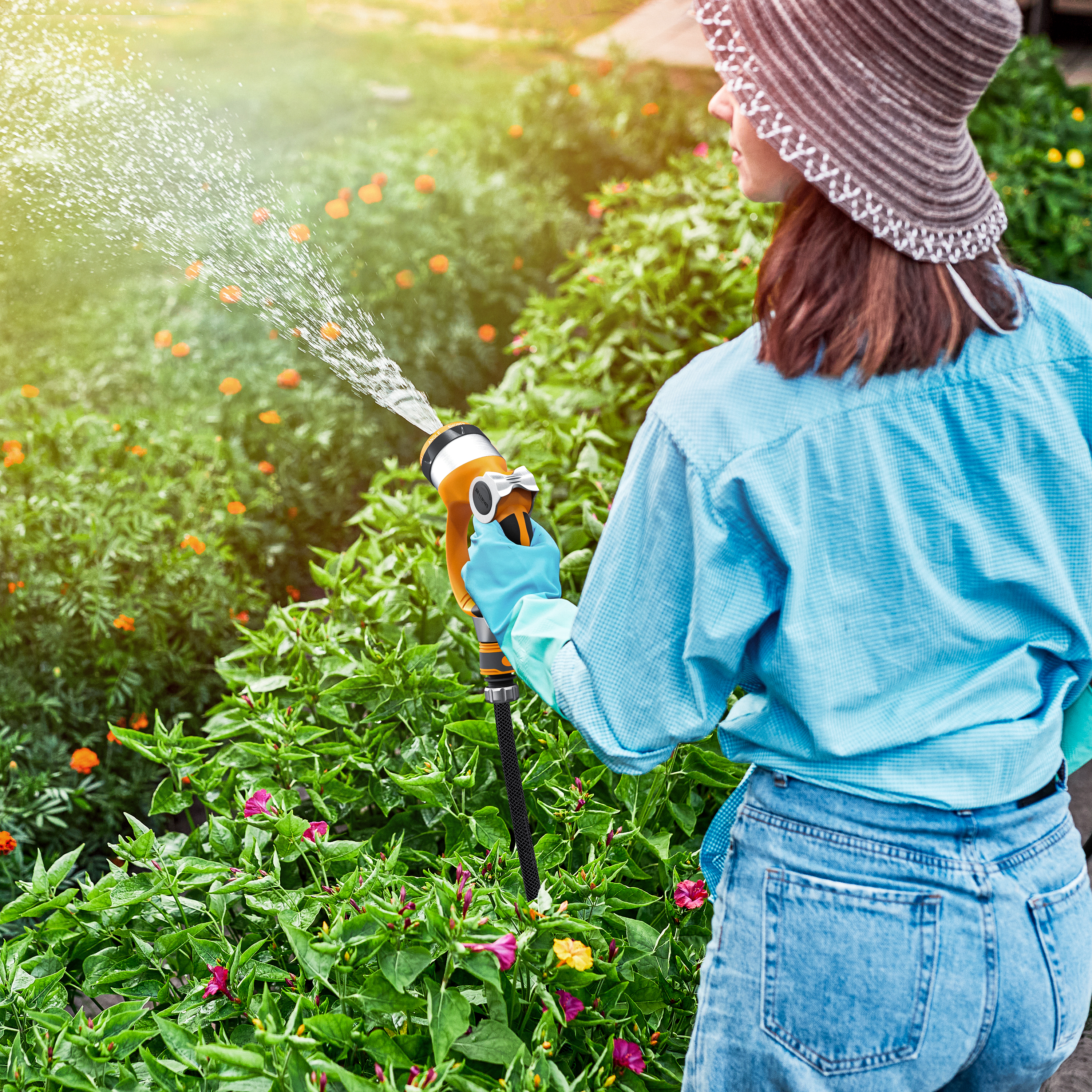 Frau mit Hut gießt grüne Pflanzen im Garten mit einer Wasserspritze.