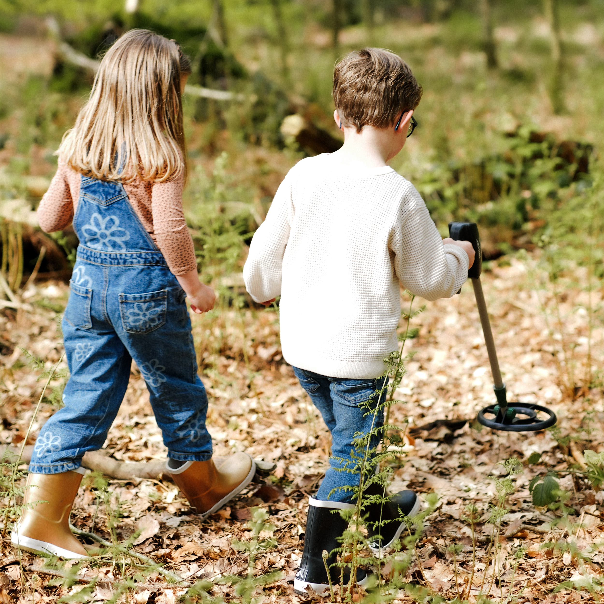 Dos niños caminan por un bosque lleno de hojas, con un niño usando un detector de metales.
