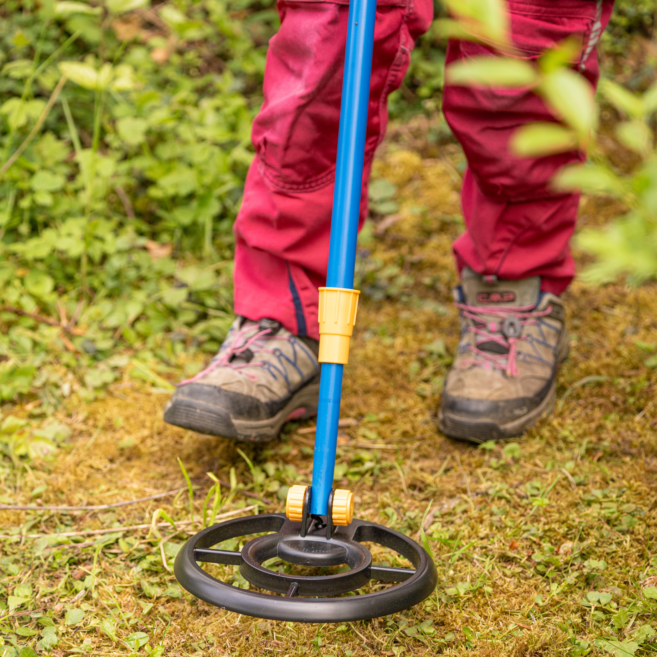 Persona con pantalones rojos y botas de montaña busca metal en un bosque cubierto de hierba con un detector azul.