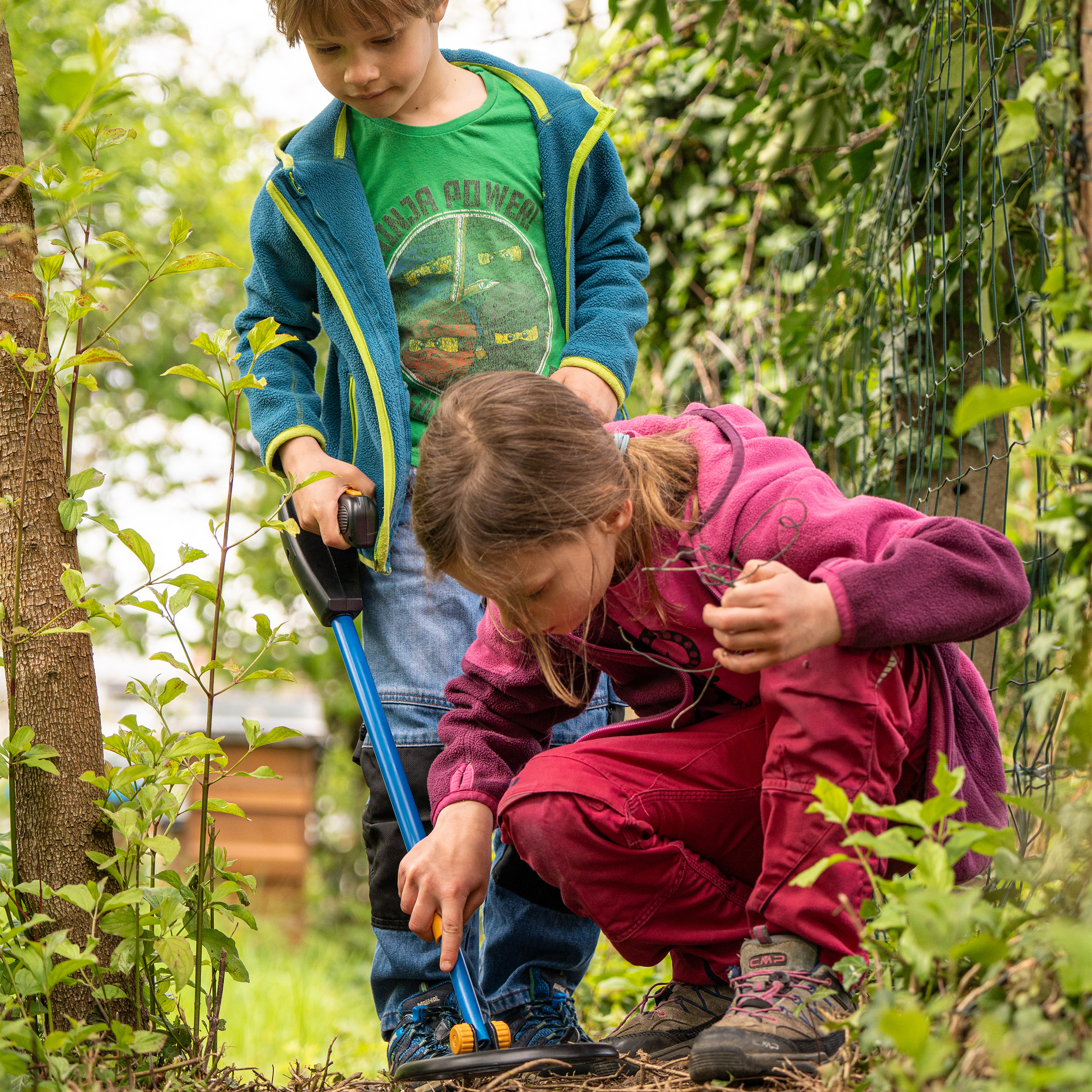 Dos niños explorando en un jardín, un niño con un detector de metales y una niña buscando en el suelo.