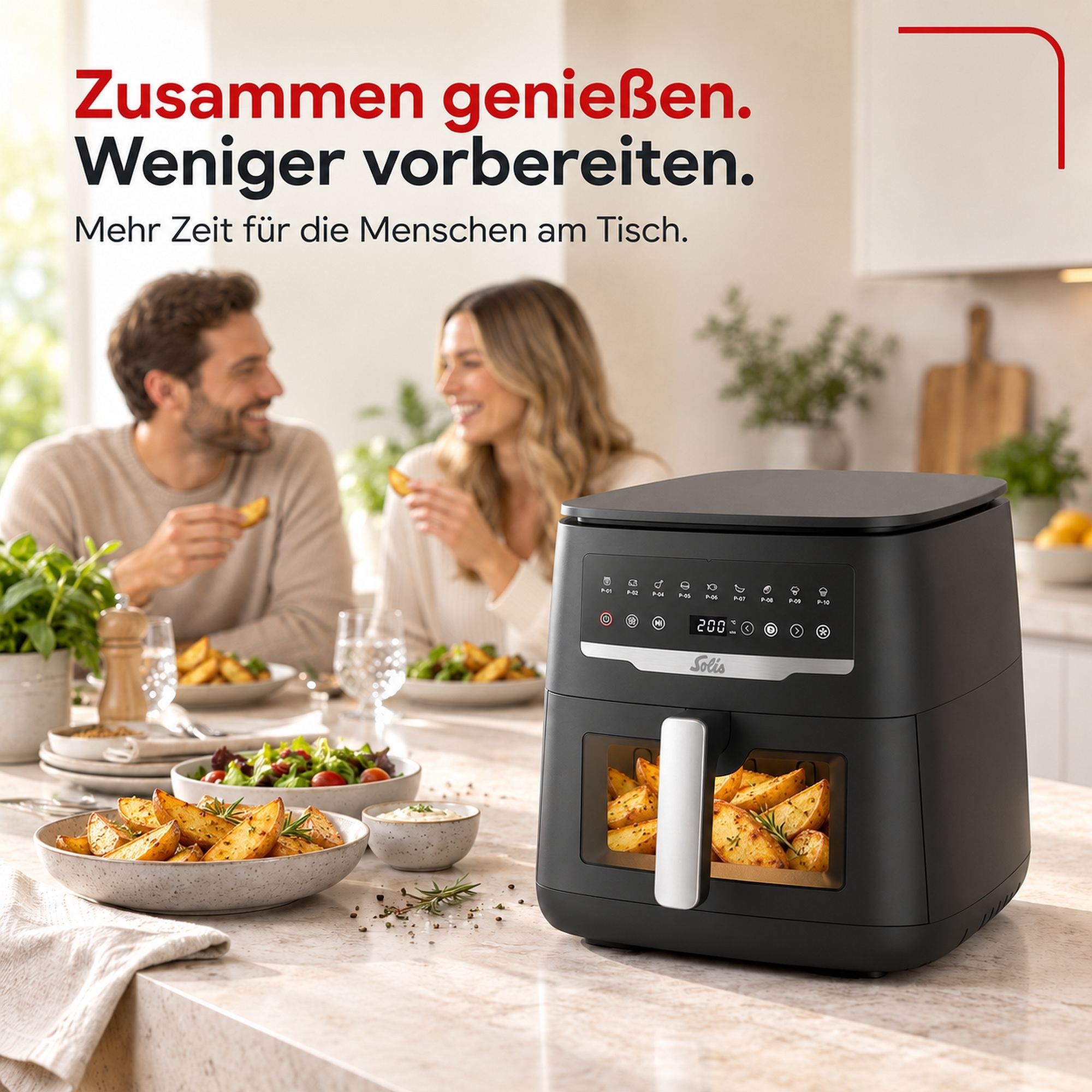 Schwarze Heißluftfritteuse auf Küchentheke mit Kartoffelecken; im Hintergrund lacht ein Paar am Esstisch.