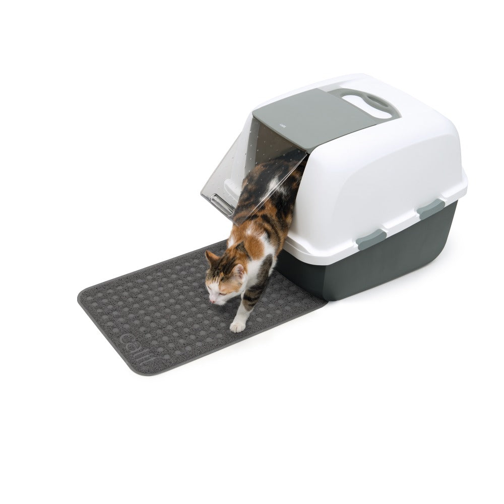 Un gato calicó sale de una caja de arena blanca y gris sobre una alfombrilla texturizada gris.