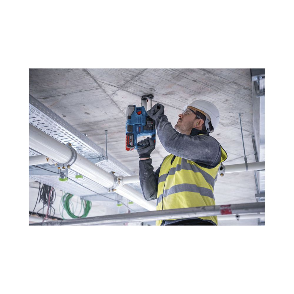 Un lavoratore con gilet ad alta visibilità e casco fora un soffitto in cemento con un trapano blu.