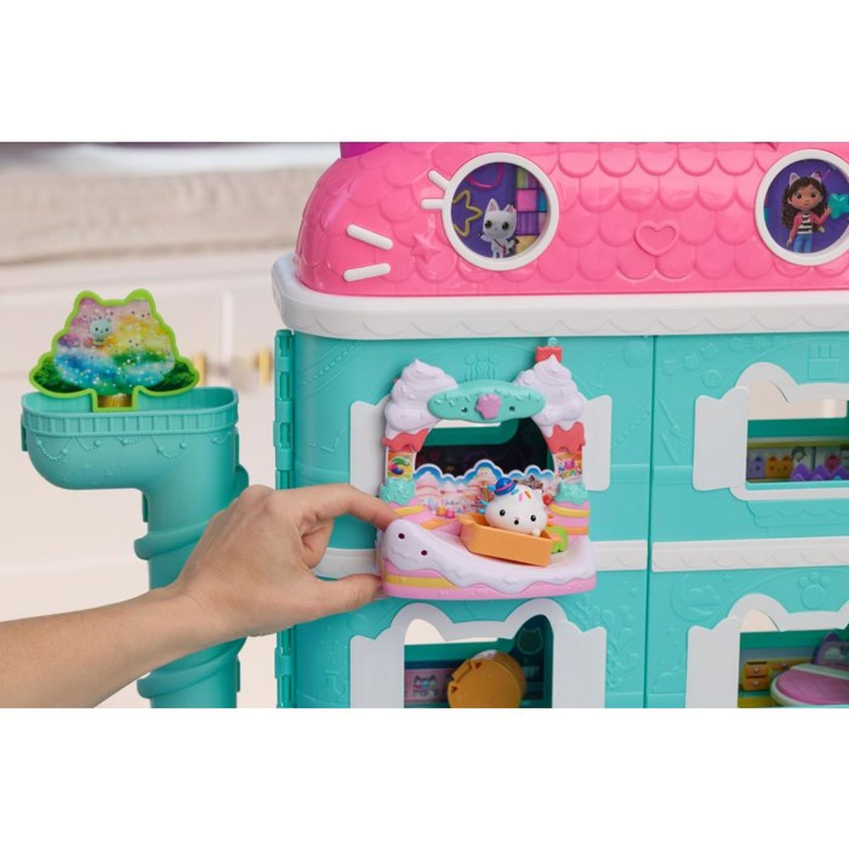 Eine Hand bewegt einen Aufzug mit einer kleinen weißen Katzenfigur an einem türkisen und pinken Puppenhaus.