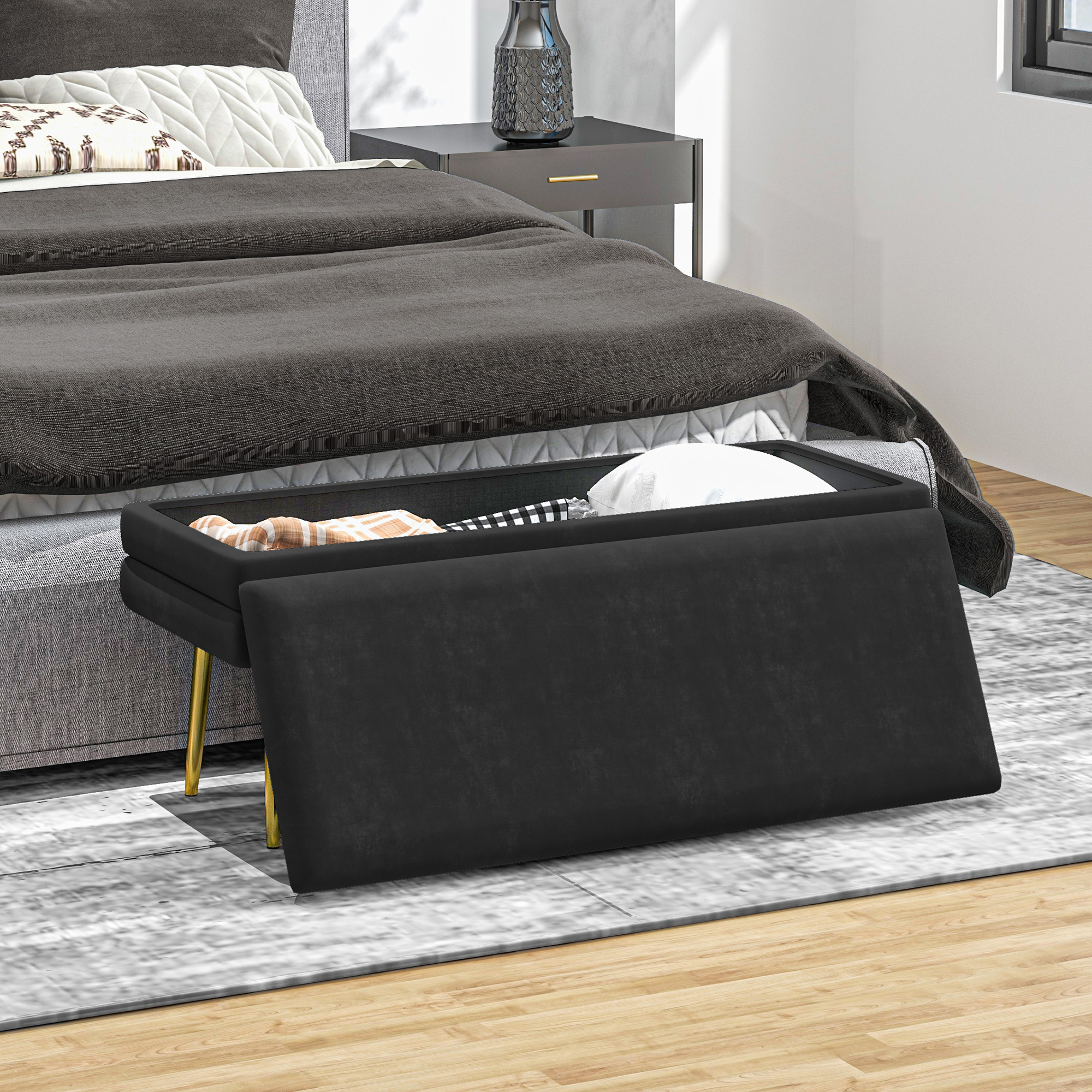 Un banco de almacenamiento negro con patas doradas frente a una cama gris en un dormitorio moderno.