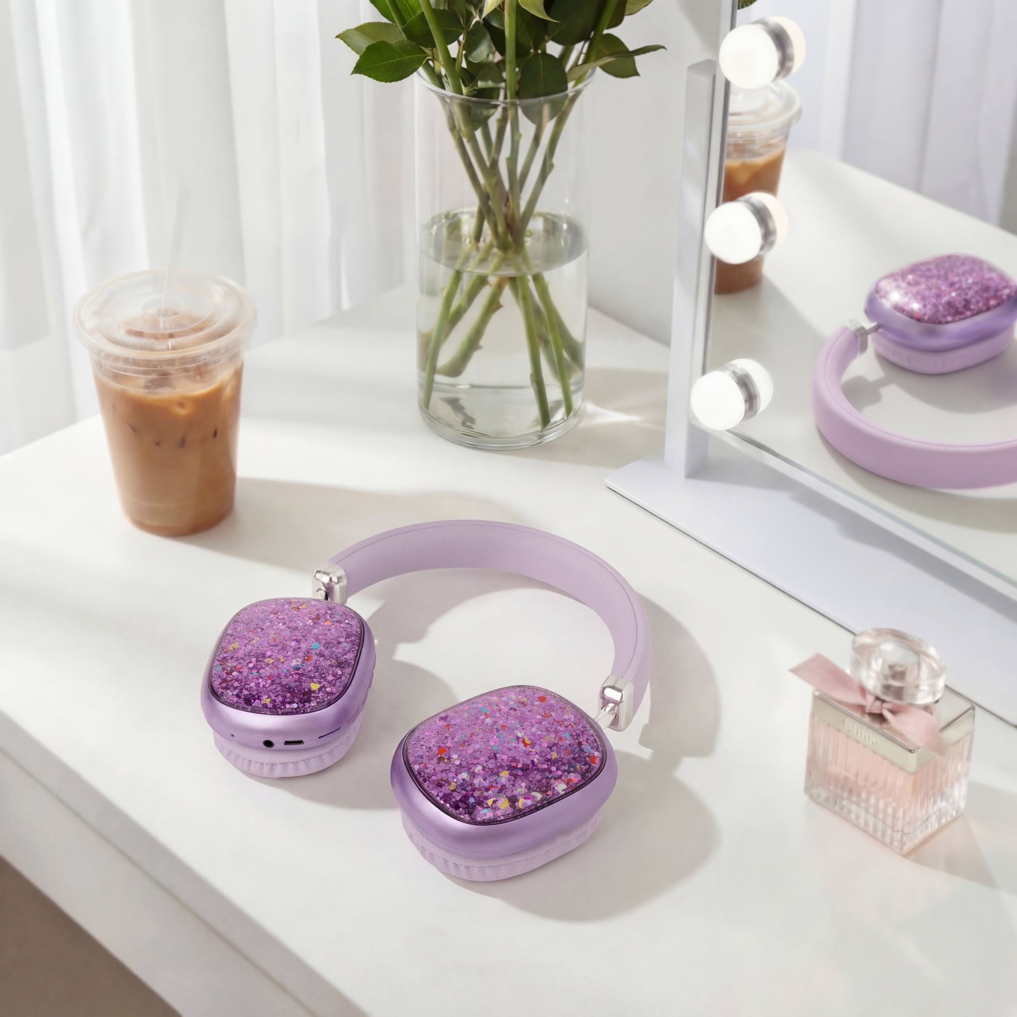Glitzernde violette Kopfhörer auf weißem Tisch mit Eiskaffee, Blumen und Parfüm.
