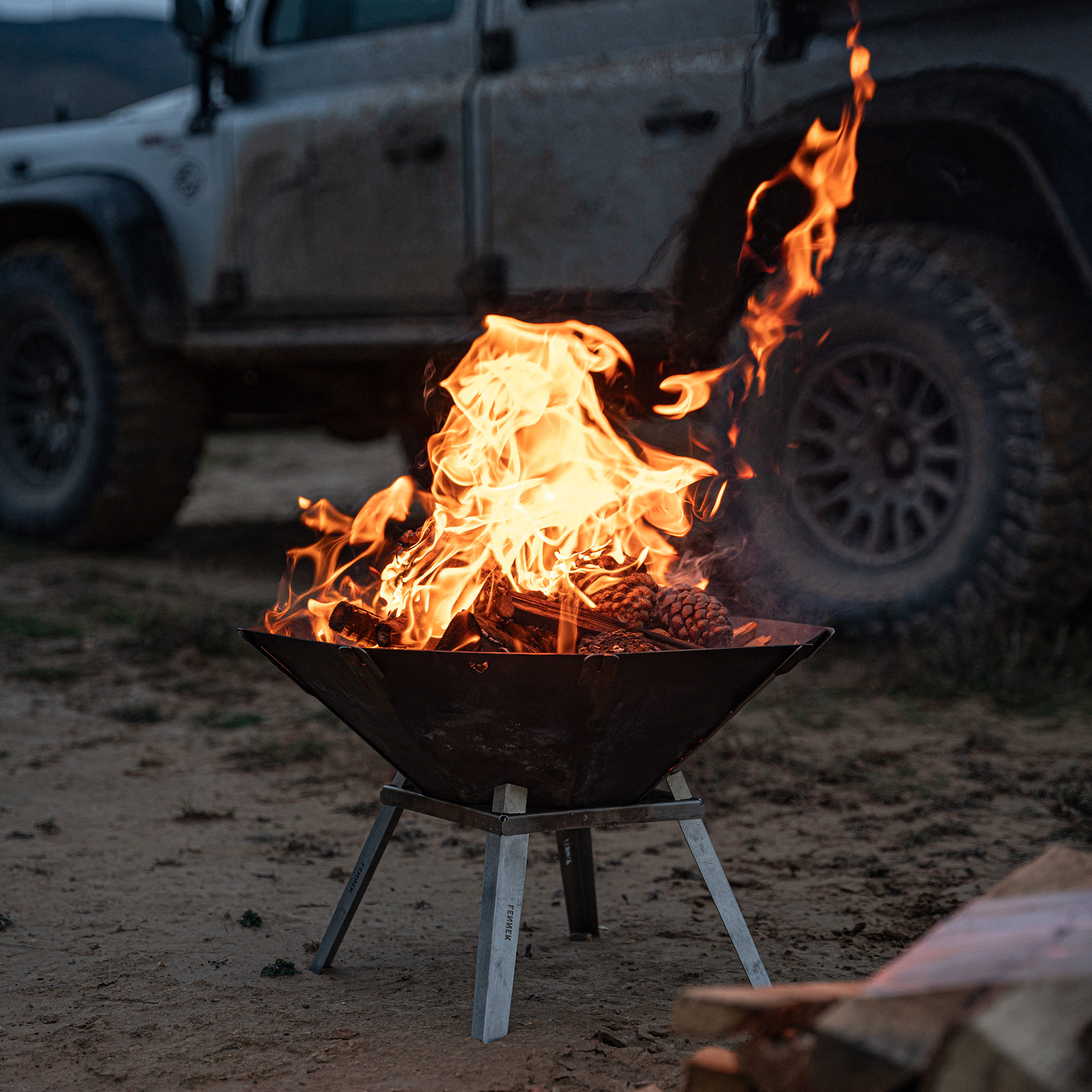 Ein Lagerfeuer brennt in einer Metallschale auf dem Boden, im Hintergrund steht ein Geländewagen.