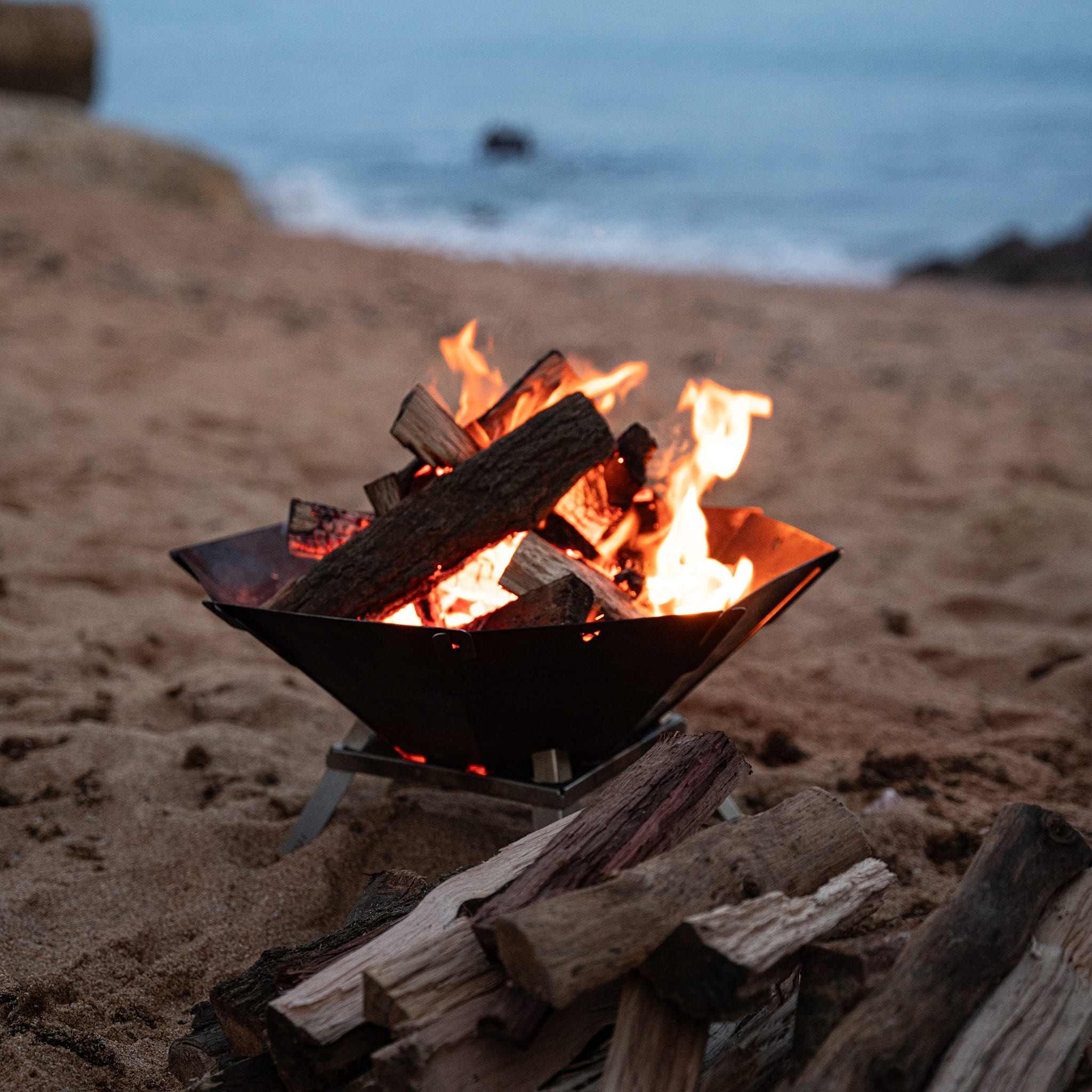 Ein Lagerfeuer brennt in einer Metallschale am sandigen Strand mit dem Meer im Hintergrund.
