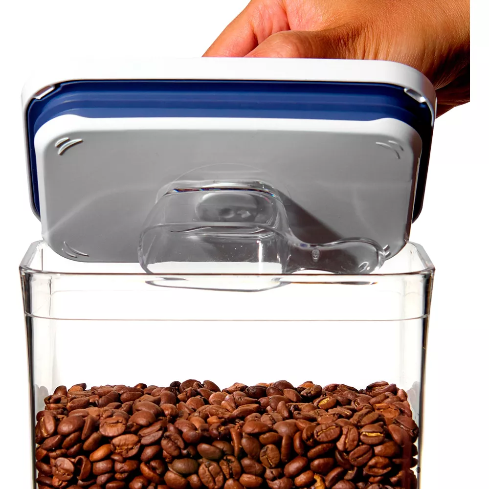 Una mano apre un contenitore trasparente pieno di chicchi di caffè, con un cucchiaio attaccato al coperchio.