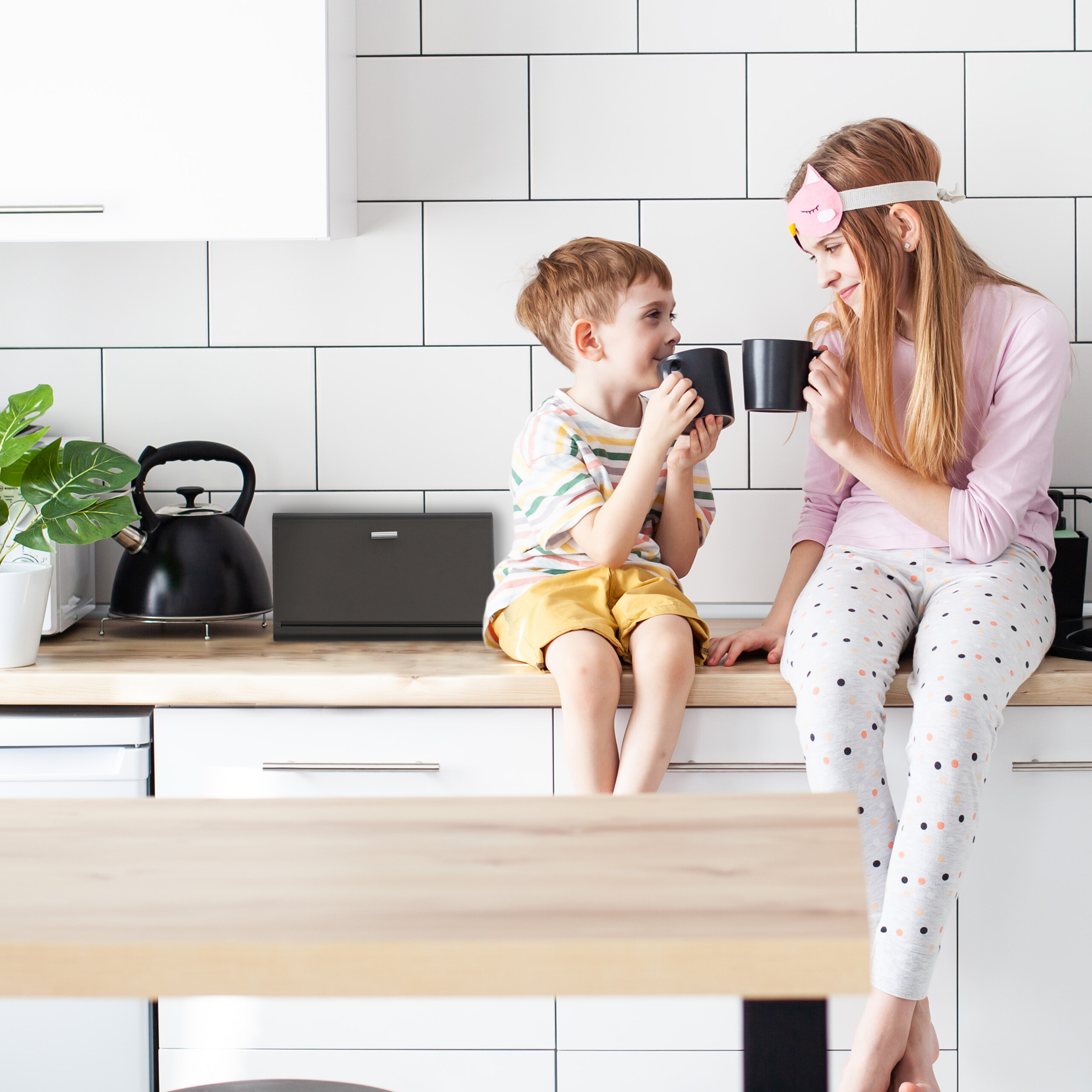 Due bambini in pigiama siedono sul bancone di una cucina bianca con tazze nere.