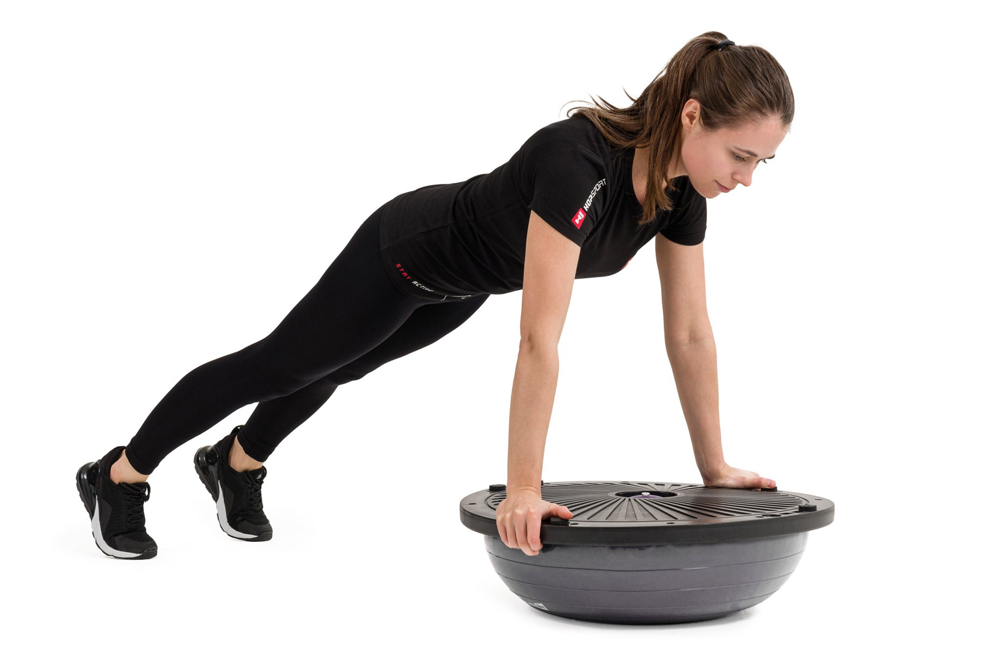 Eine Frau in schwarzem Sportoutfit macht einen Liegestütz auf einem Balance Board vor weißem Hintergrund.