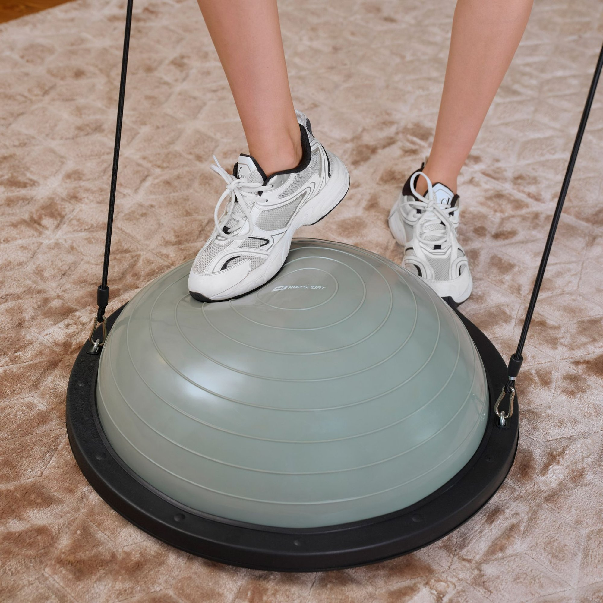 Eine Person in weißen Turnschuhen steht auf einem grauen Balance-Trainer mit Widerstandsbändern auf einem Teppich.
