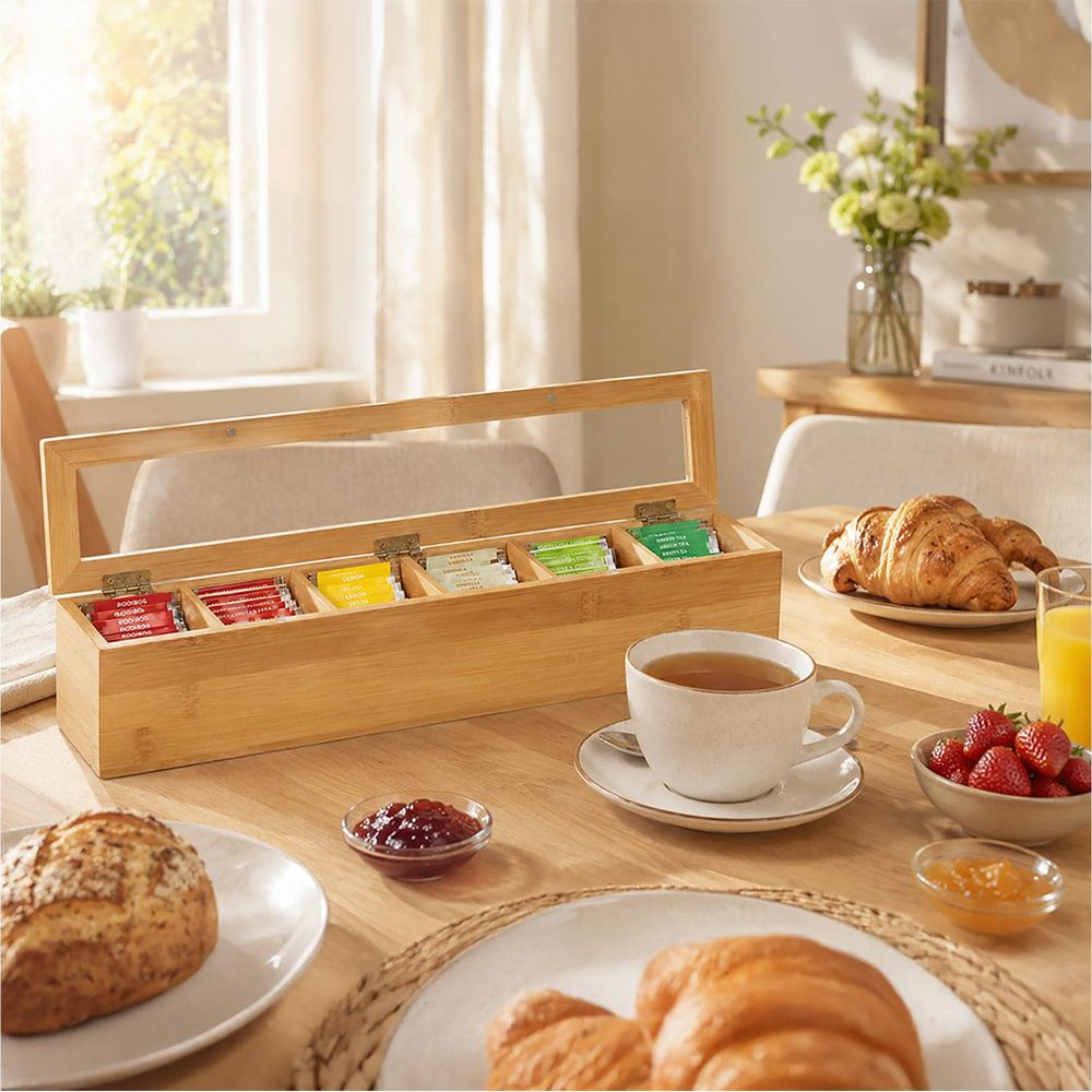 Un porta bustine di tè in legno con bustine su un tavolo vicino alla colazione con croissant, caffè e marmellata.