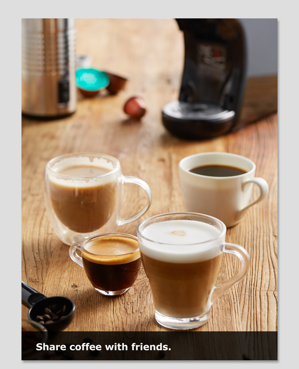 Quattro tazze di varie bevande a base di caffè su un tavolo di legno con una macchina da caffè sullo sfondo.