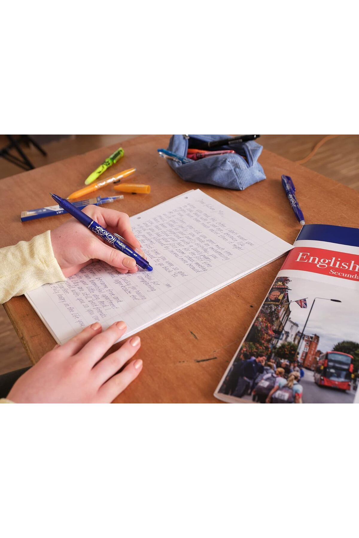 Bir kişi ahşap masada çizgili bir deftere mavi kalemle yazıyor, etrafında kalemler ve bir kitap var.