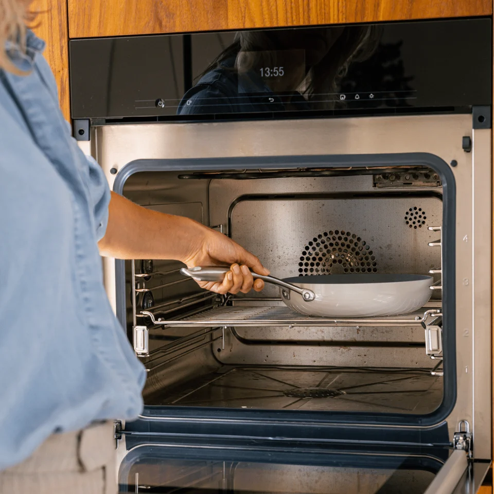 A person places a frying pan inside an open, modern stainless steel oven in a kitchen.