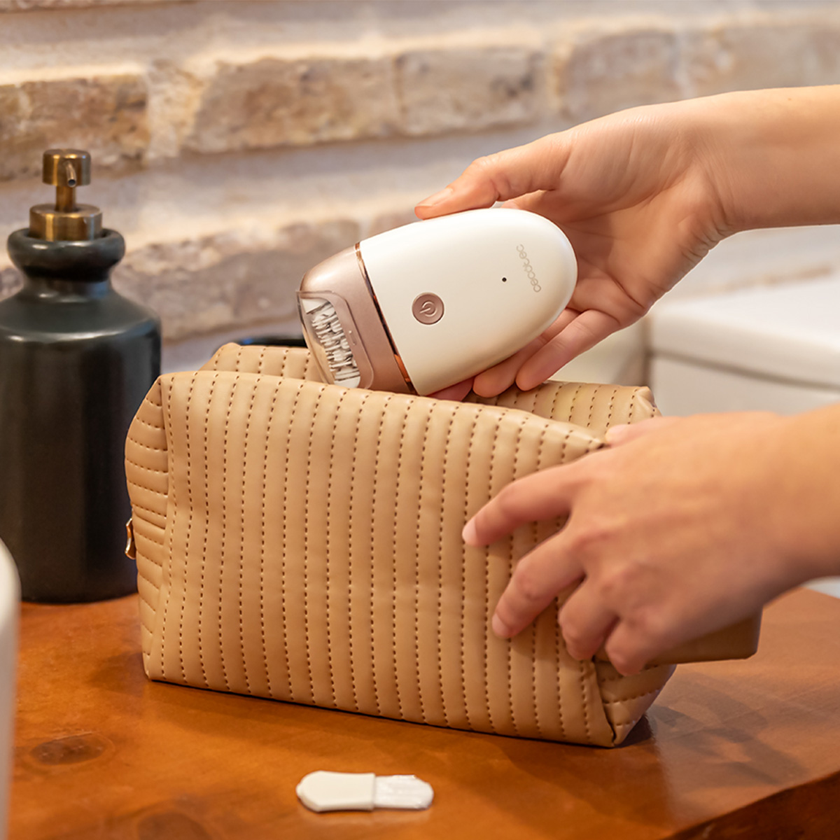 Una persona coloca una depiladora en un neceser beige sobre una mesa de madera.