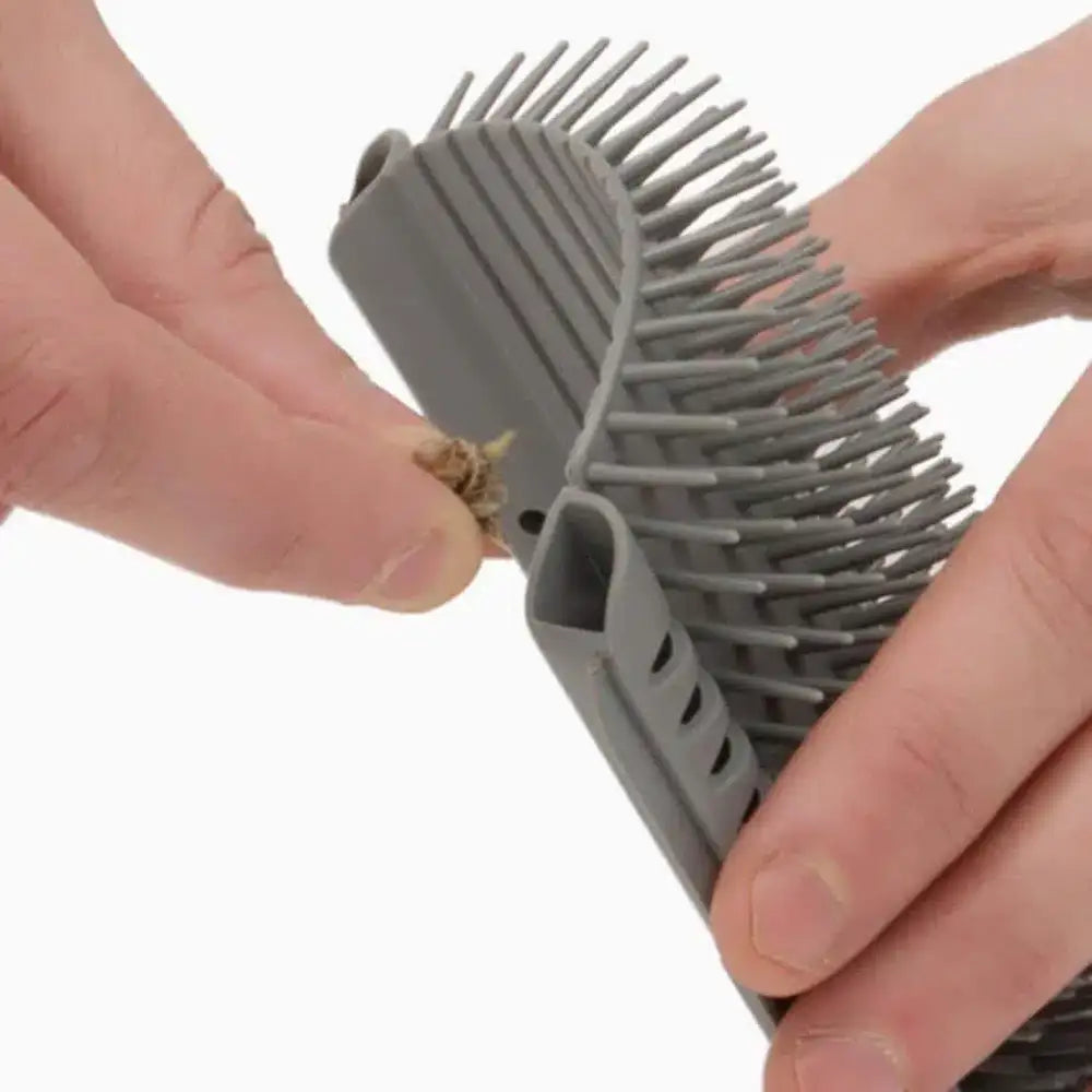 Manos sostienen un cepillo para mascotas de silicona gris curvado, quitando pelo atrapado de las cerdas de goma.