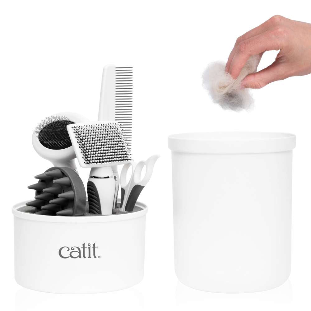 Una mano tira pelo en un cubo blanco Catit junto a un recipiente con herramientas de aseo para gatos.
