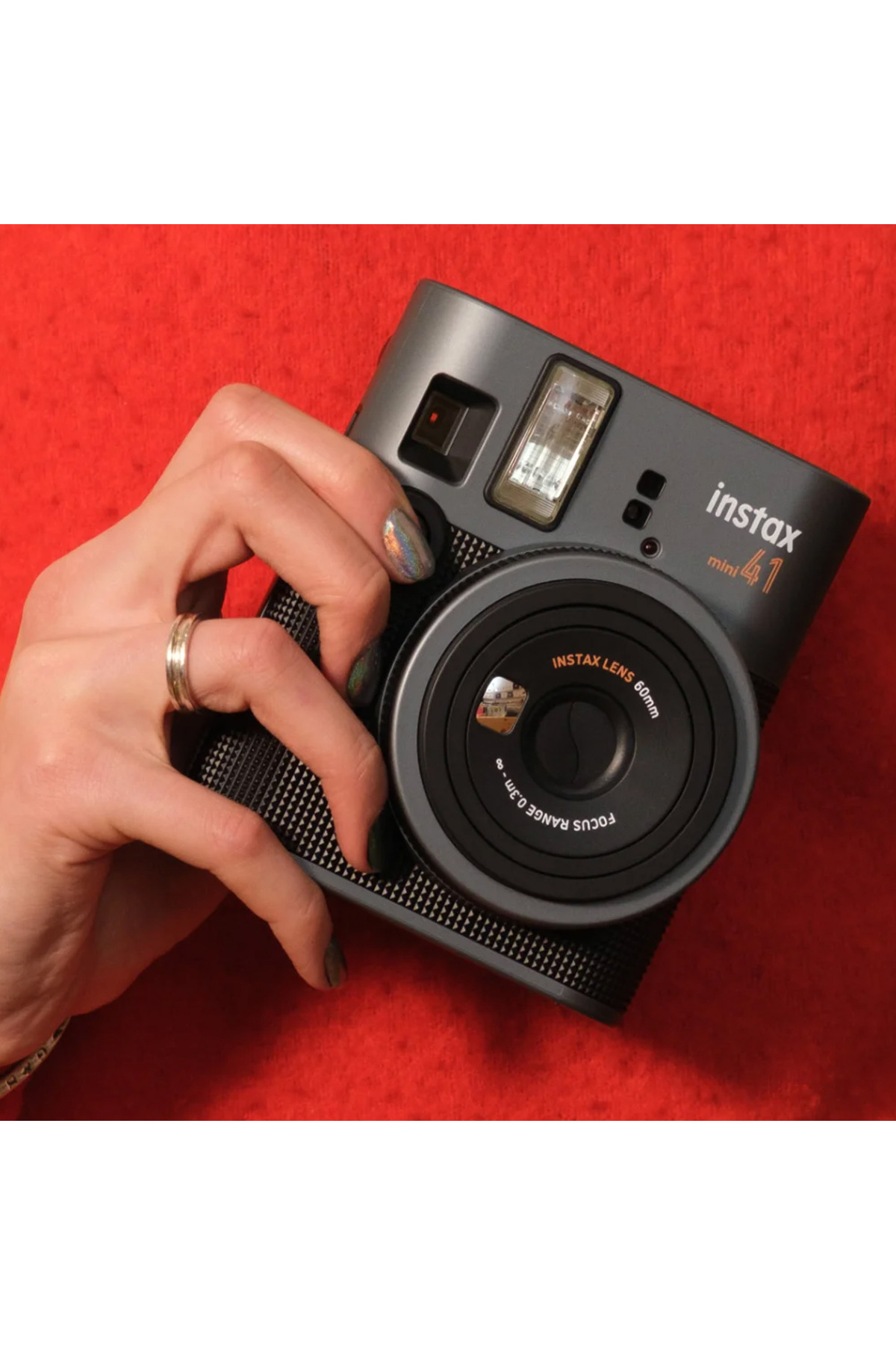 Kırmızı dokulu bir arka plan önünde koyu gri bir Instax mini kamera tutan bir el.