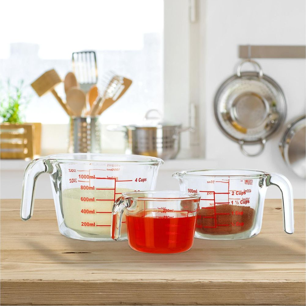Drei Glas-Messbecher mit roter Skala auf einem Holztisch, im Hintergrund eine Küche mit Utensilien.