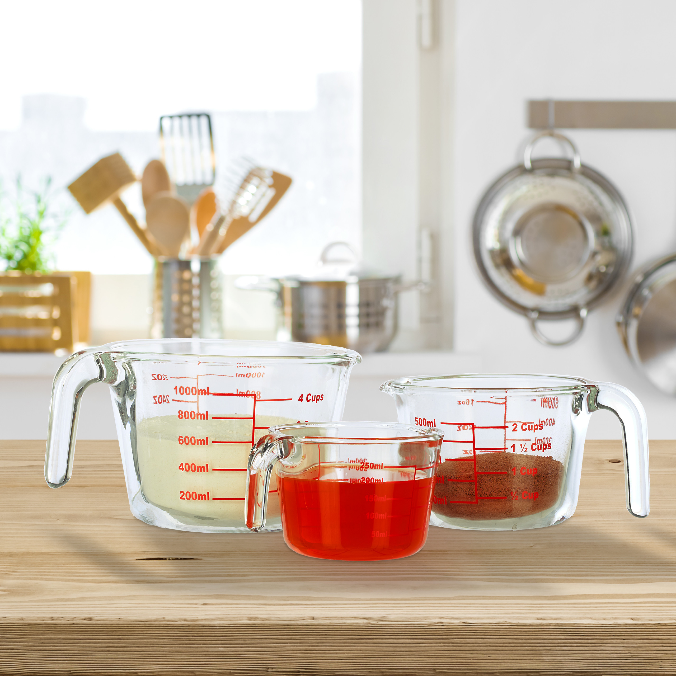 Drei Glas-Messbecher mit gelbem Pulver, roter Flüssigkeit und braunem Pulver stehen auf einem Holztisch in einer Küche.
