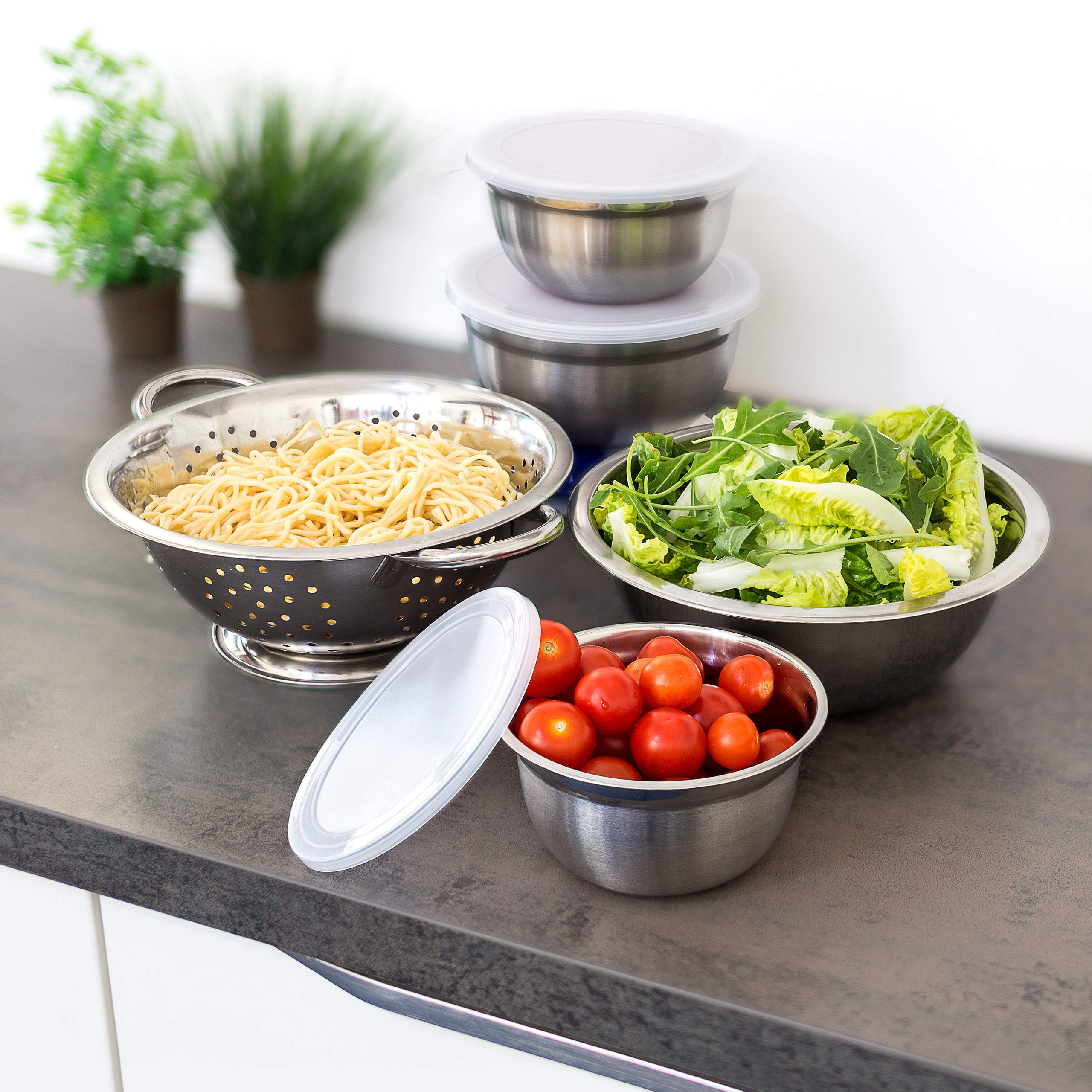 Edelstahlschüsseln und ein Sieb mit Nudeln, Salat und Tomaten auf einer Küchentheke.