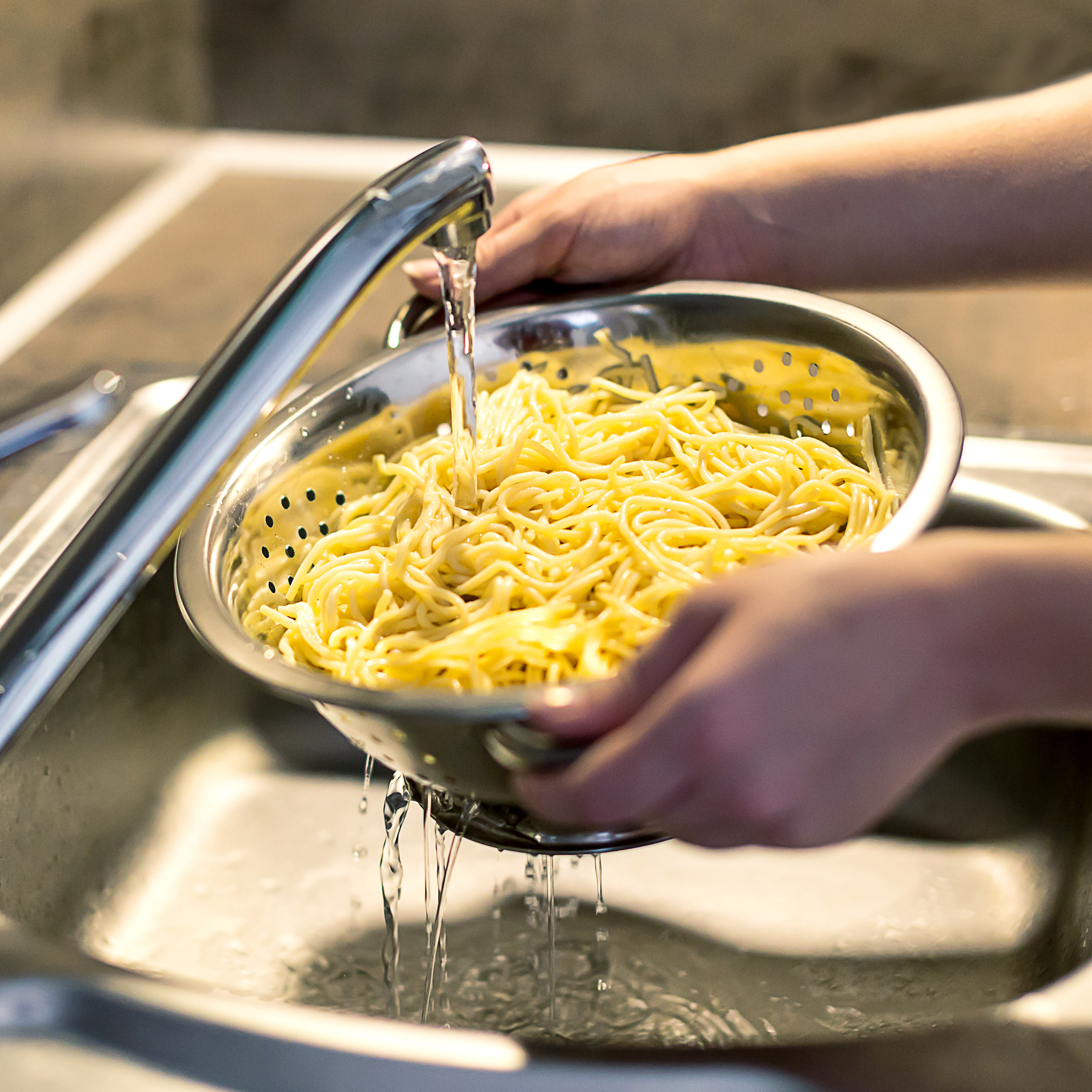 Person wäscht gekochte Spaghetti in einem Metallsieb unter fließendem Wasser über der Spüle.