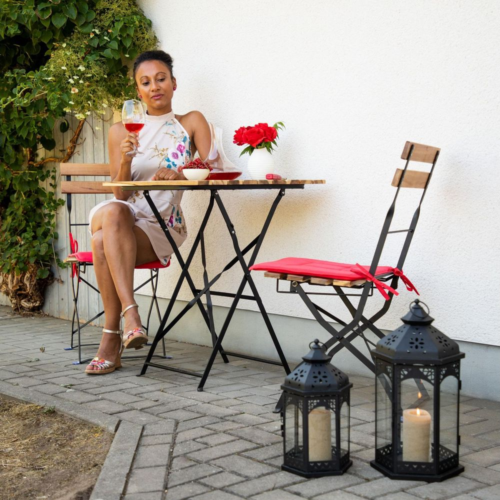 Eine Frau sitzt an einem Gartentisch, hält ein Glas Wein neben roten Blumen und zwei Laternen.